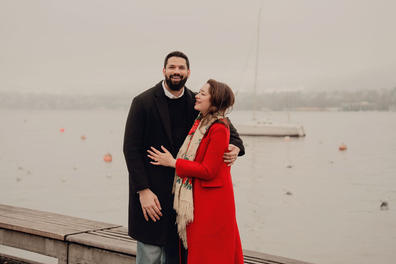 Smiling couple standing by a foggy lake, woman in red coat leaning on man in black overcoat.