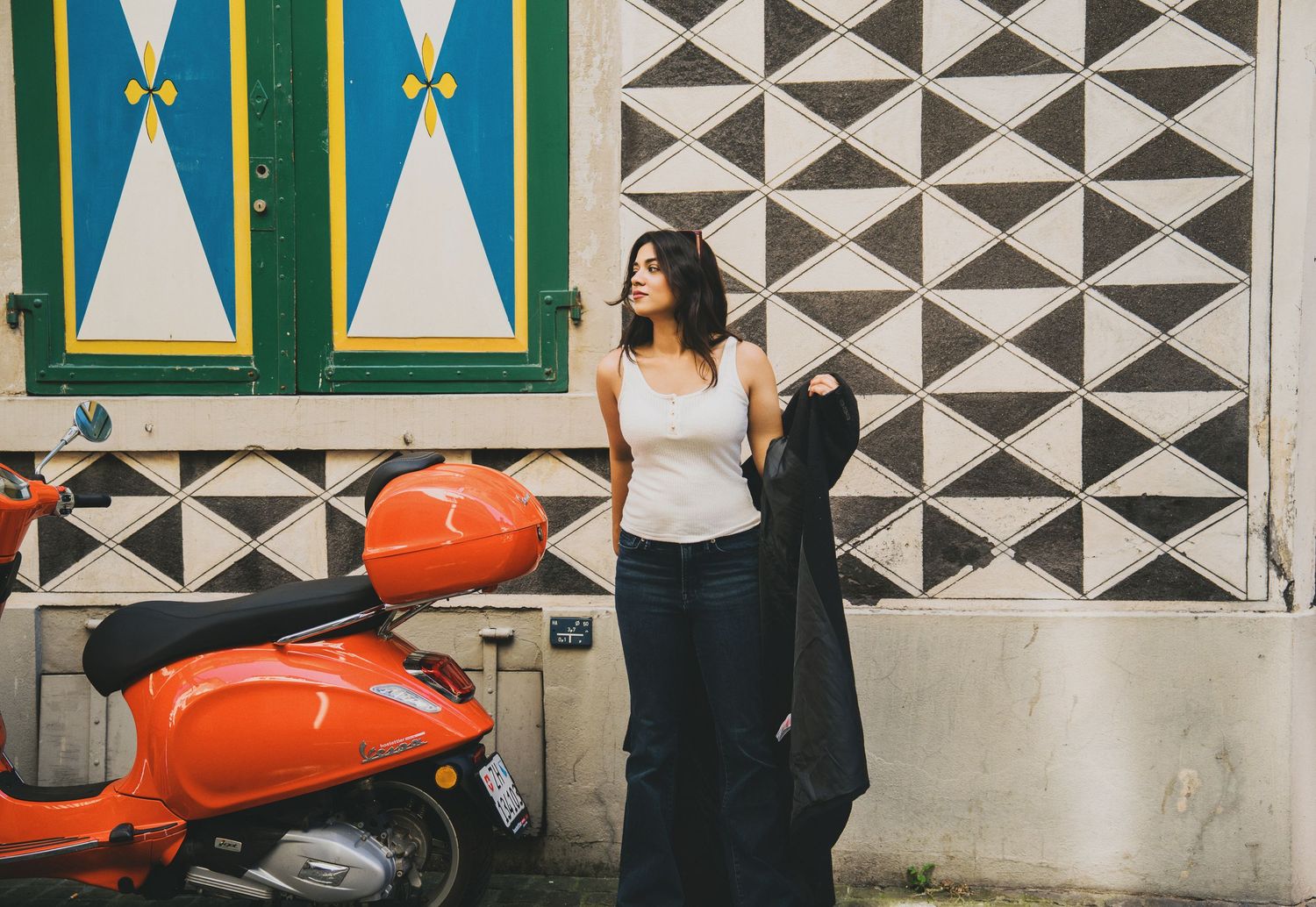 Stylish woman in white top and jeans standing beside orange scooter against geometric patterned wall.