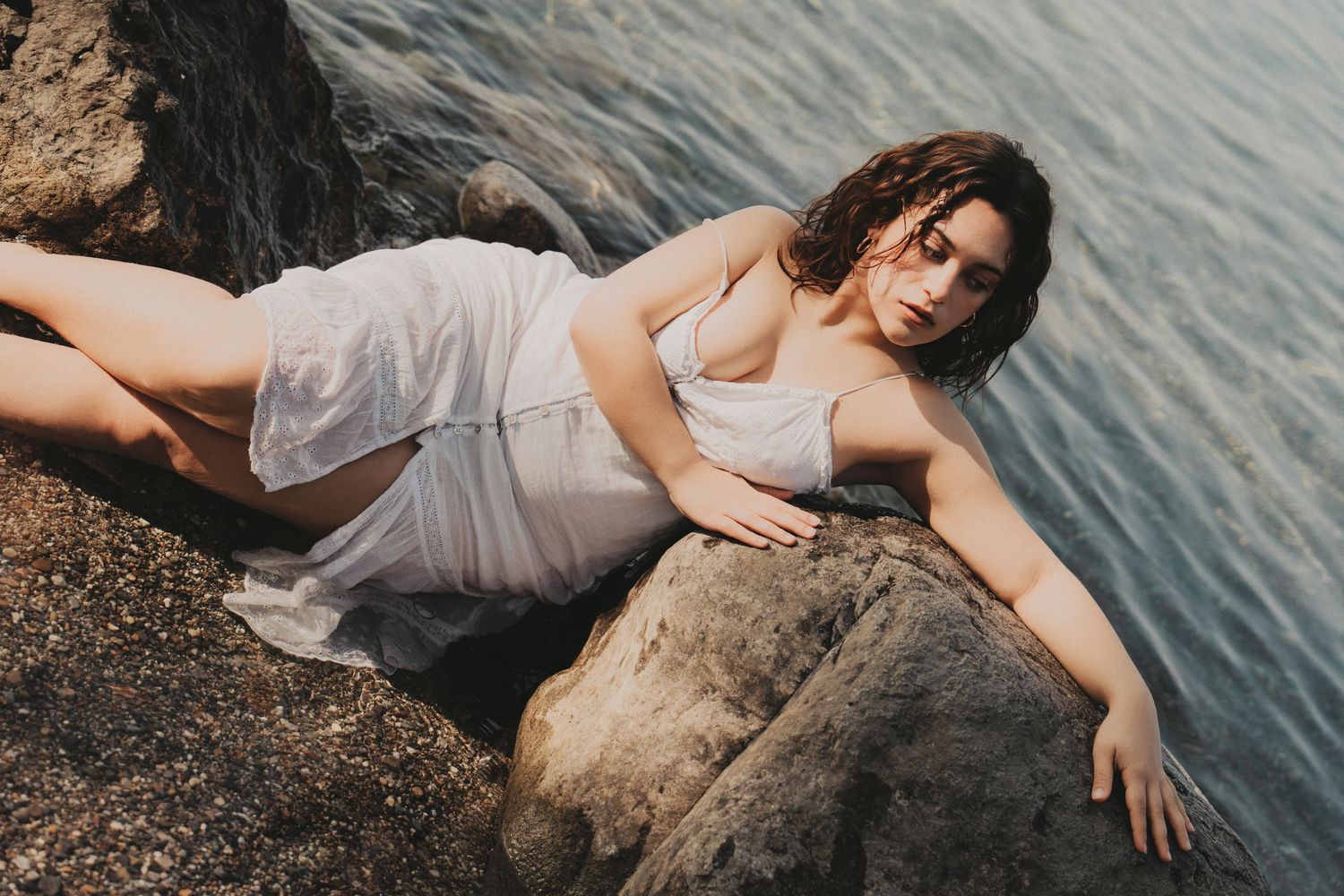 Woman in wet white dress reclining gracefully on rocky coastal cliffs by blue water.