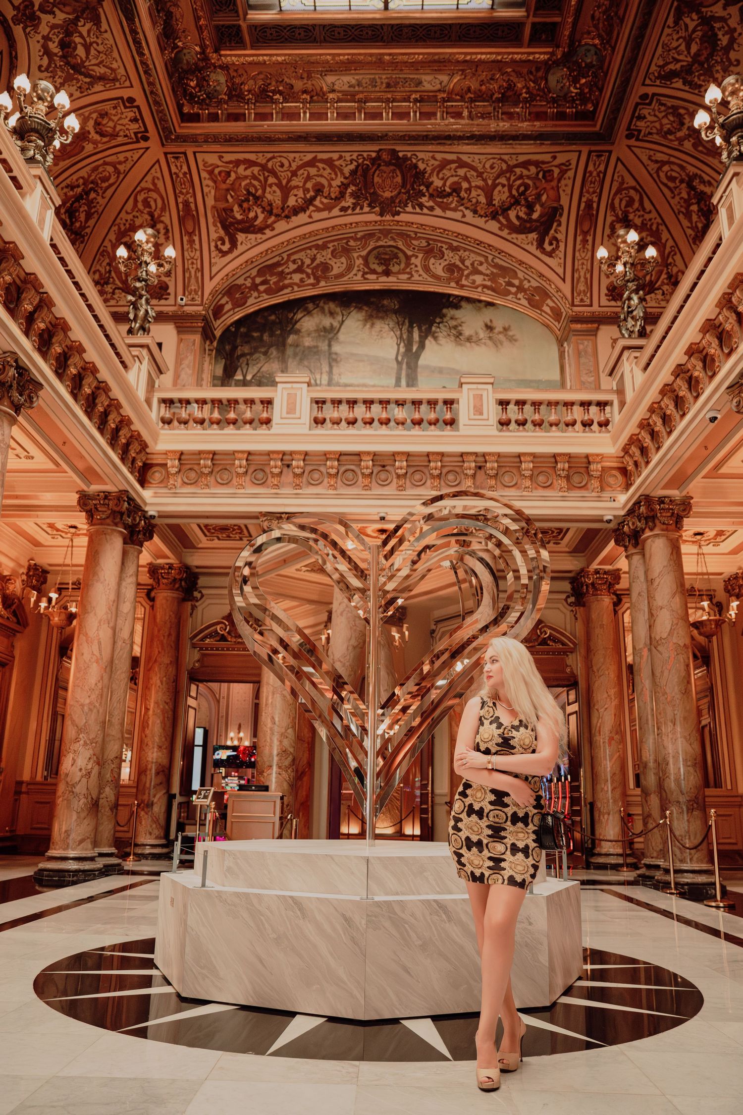 Blonde woman in black and gold dress posing beside a large golden heart art installation in a grand hall.