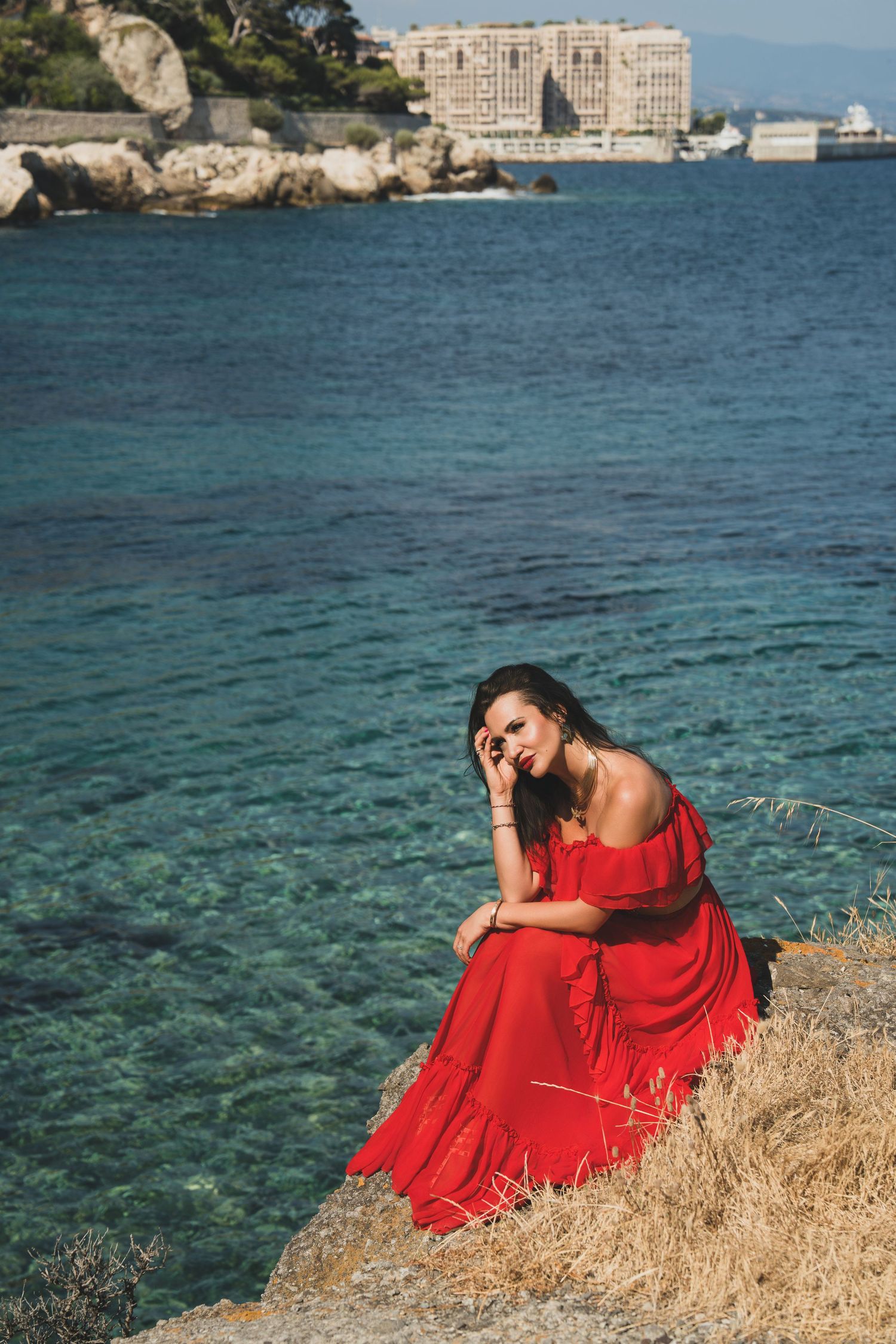 Brunette woman in red maxi dress posed on seaside rocks with hotel in background.