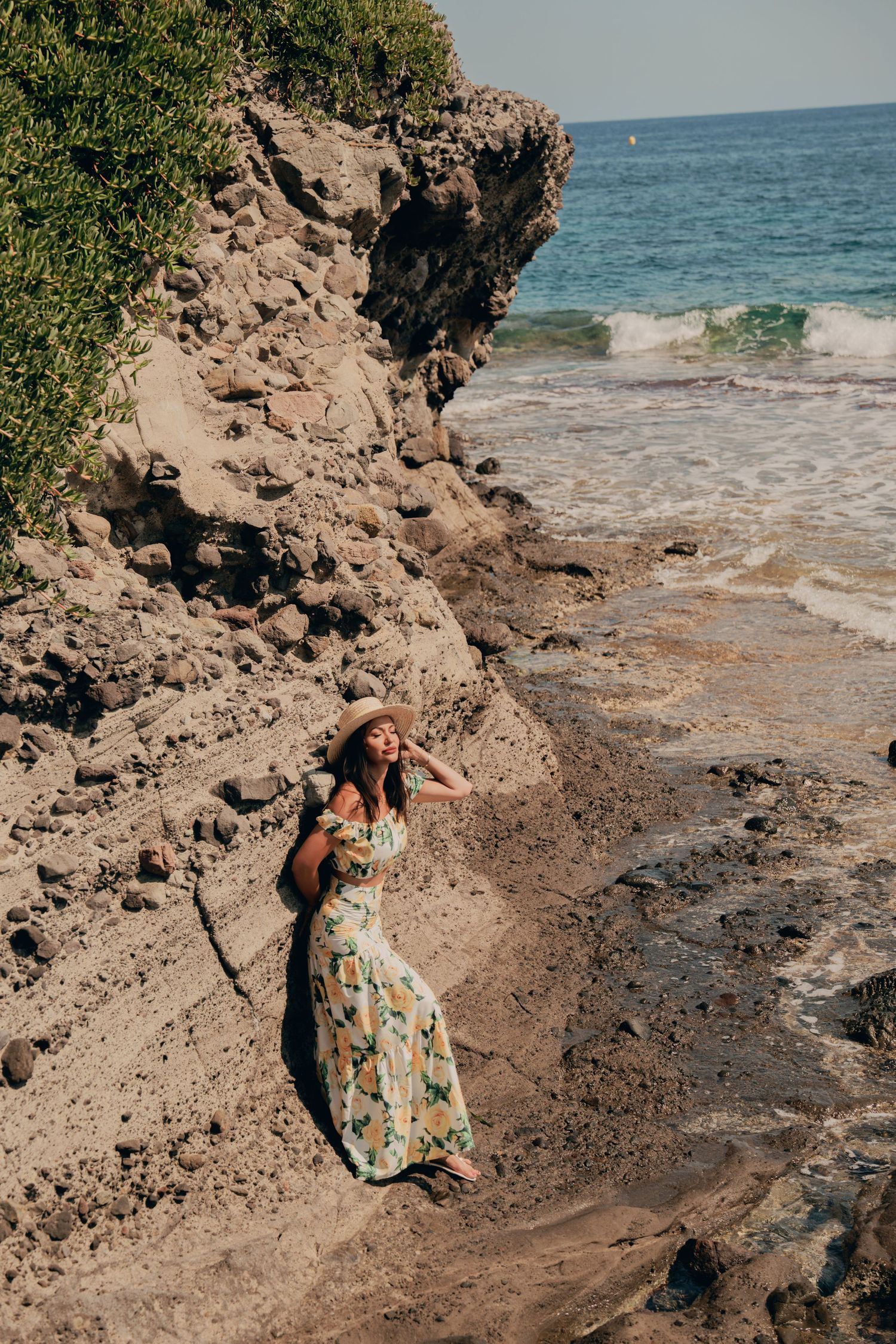 Woman in floral dress and sun hat sitting on sandy rocks by the ocean shore.