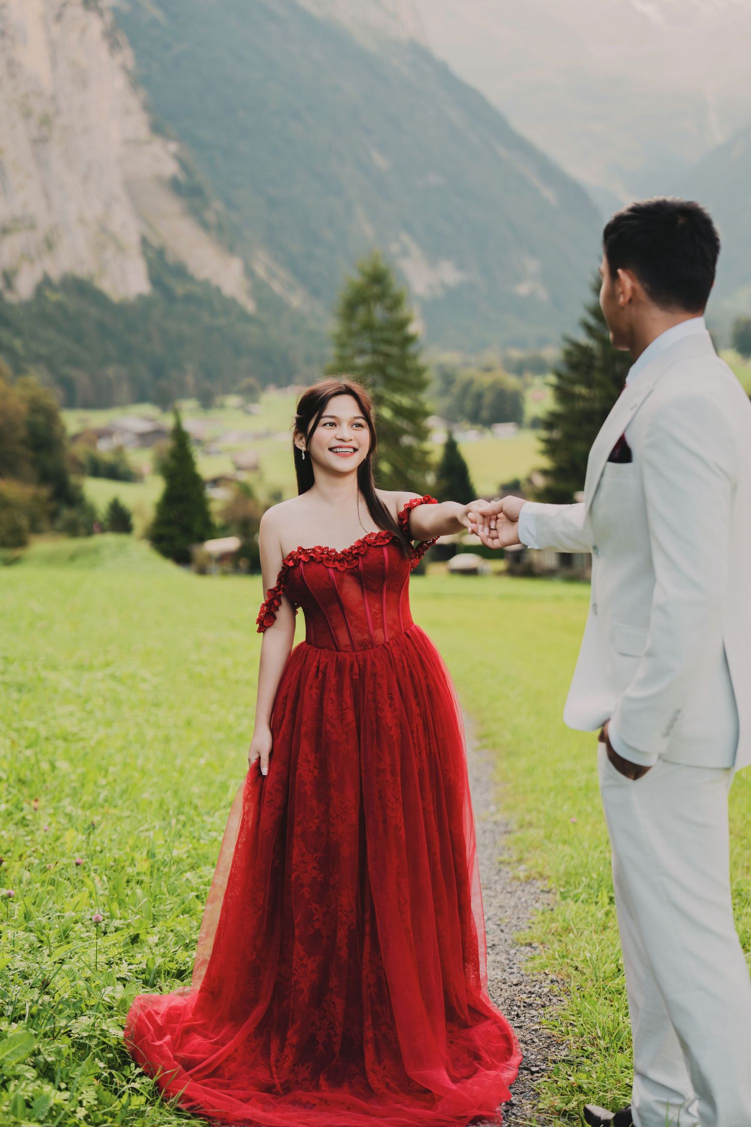 Laughing bride in red off-shoulder gown holds groom's hand in Swiss alpine valley.