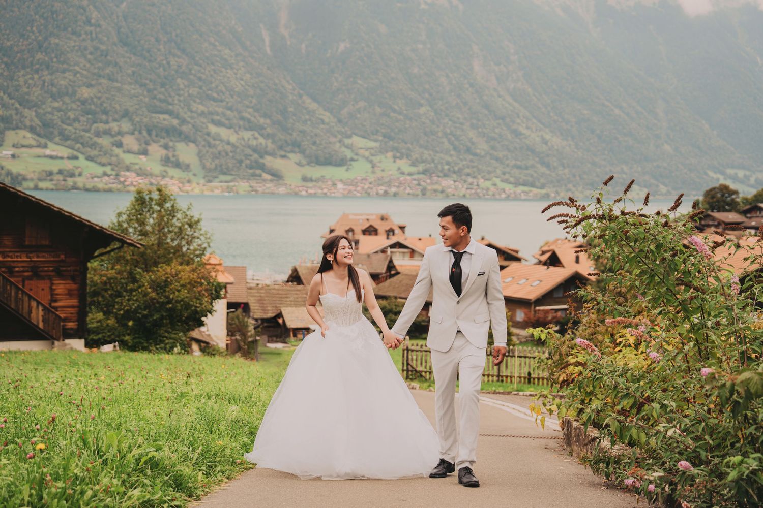 Bride and groom walking hand in hand through Swiss alpine village with lake and mountains behind.