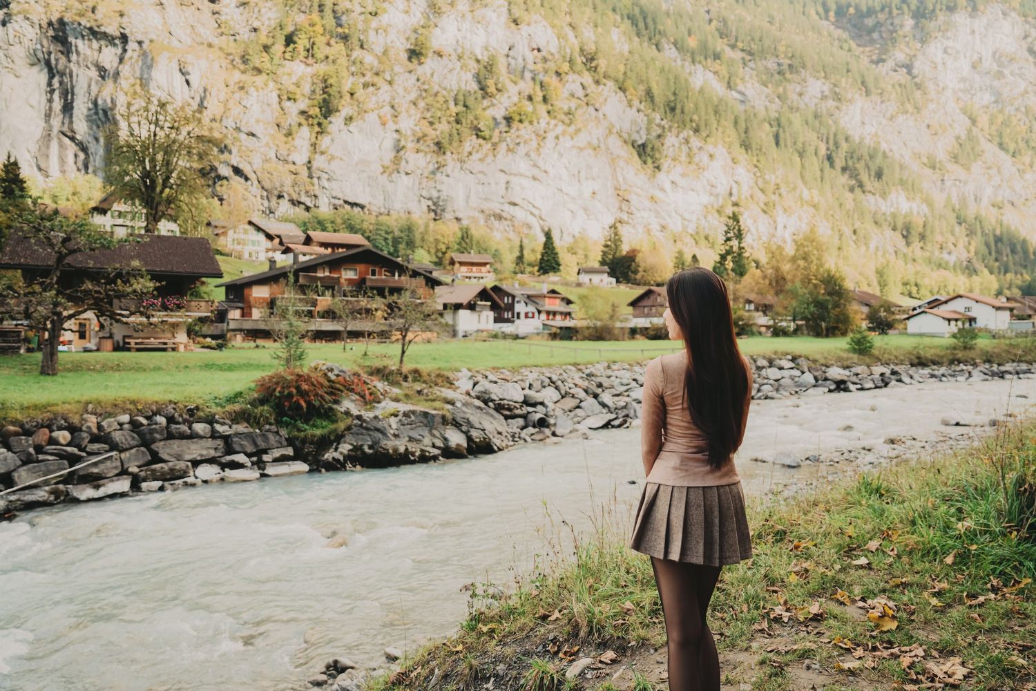 Woman with long dark hair standing by a glacial river admiring a picturesque Swiss mountain village in autumn.
