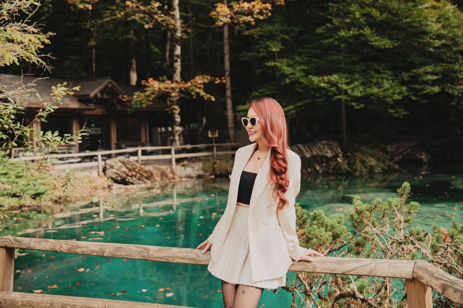 Young woman with long red hair in white blazer and sunglasses standing by turquoise alpine lake in forest.
