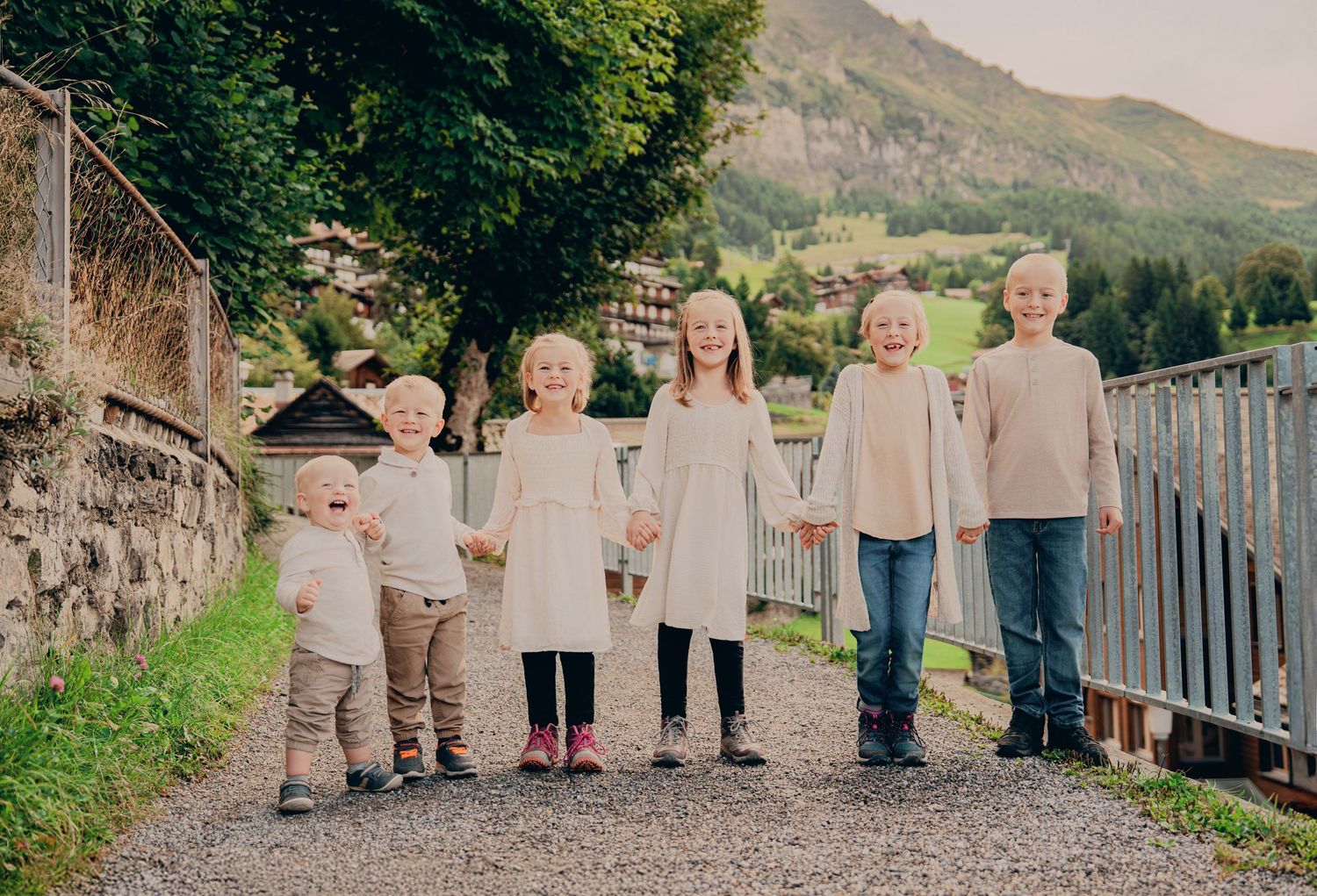 Six young children hold hands while standing on a gravel path with mountain scenery behind them.
