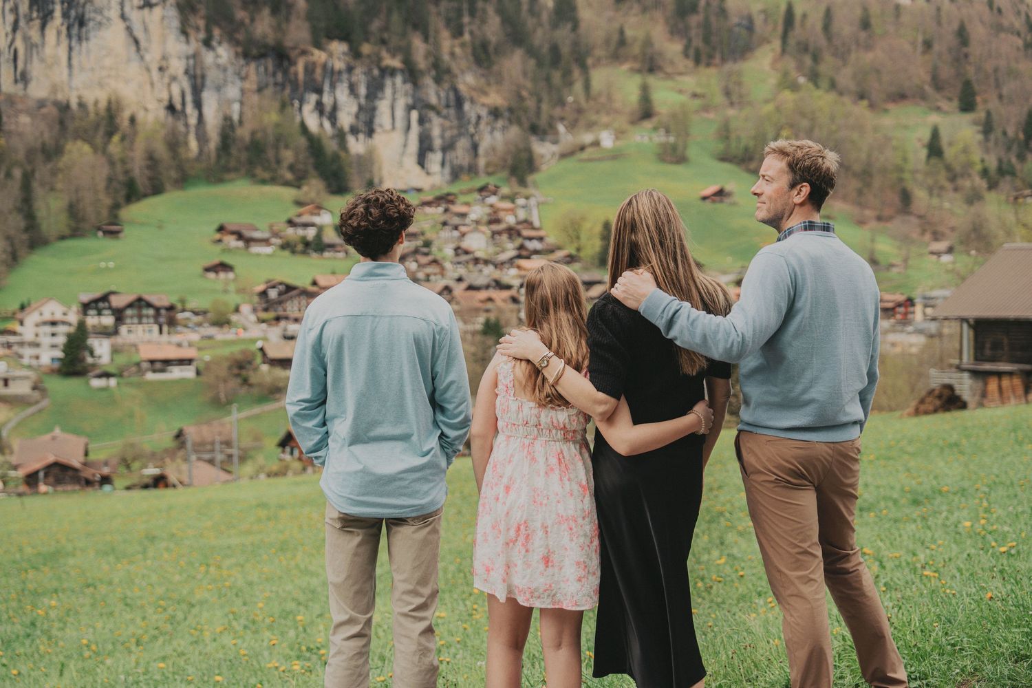 Family of four viewed from behind admiring picturesque Swiss alpine village on green hillside.