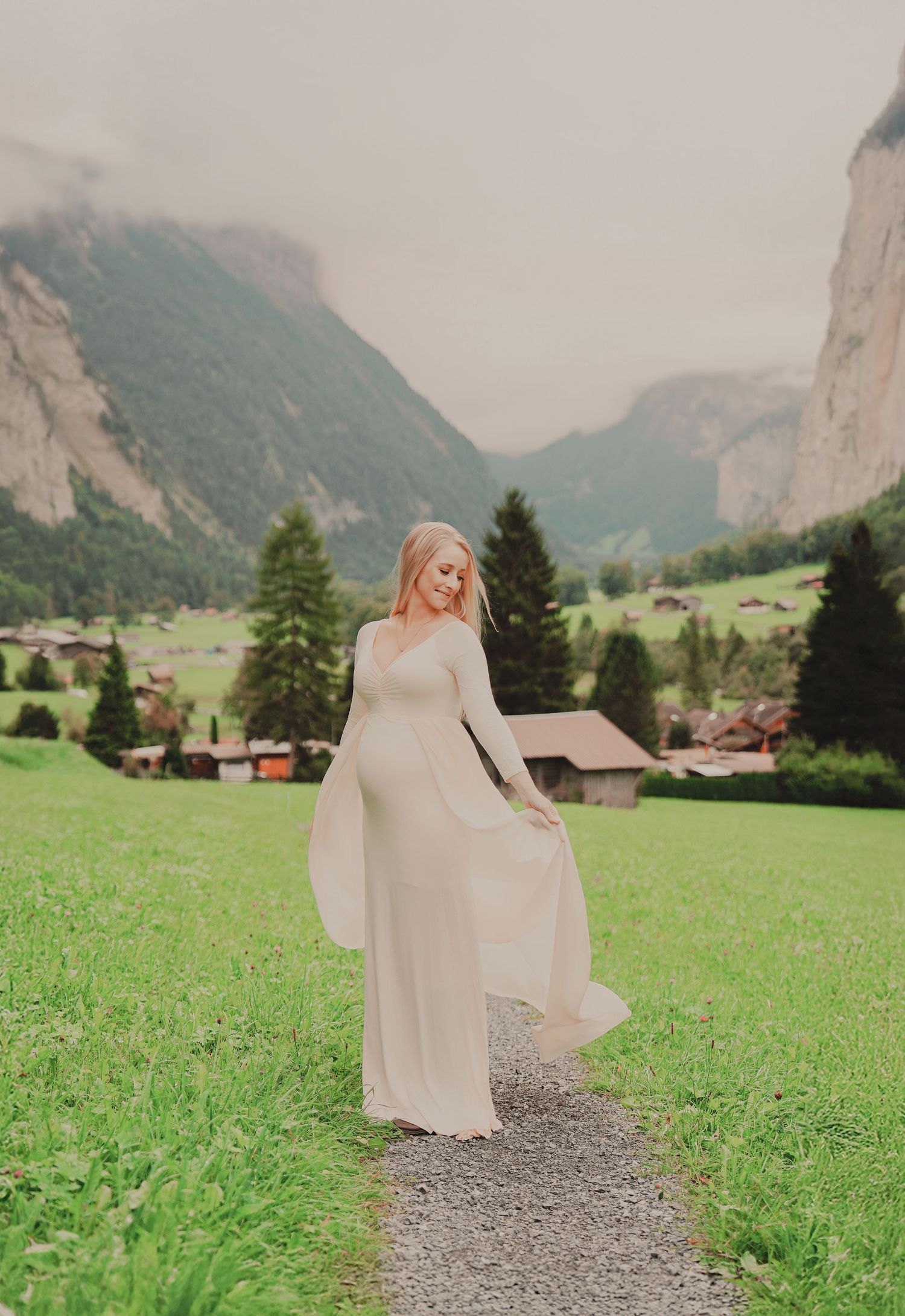 Smiling pregnant woman in flowing white maternity gown twirling on a path in a stunning Swiss mountain valley.
