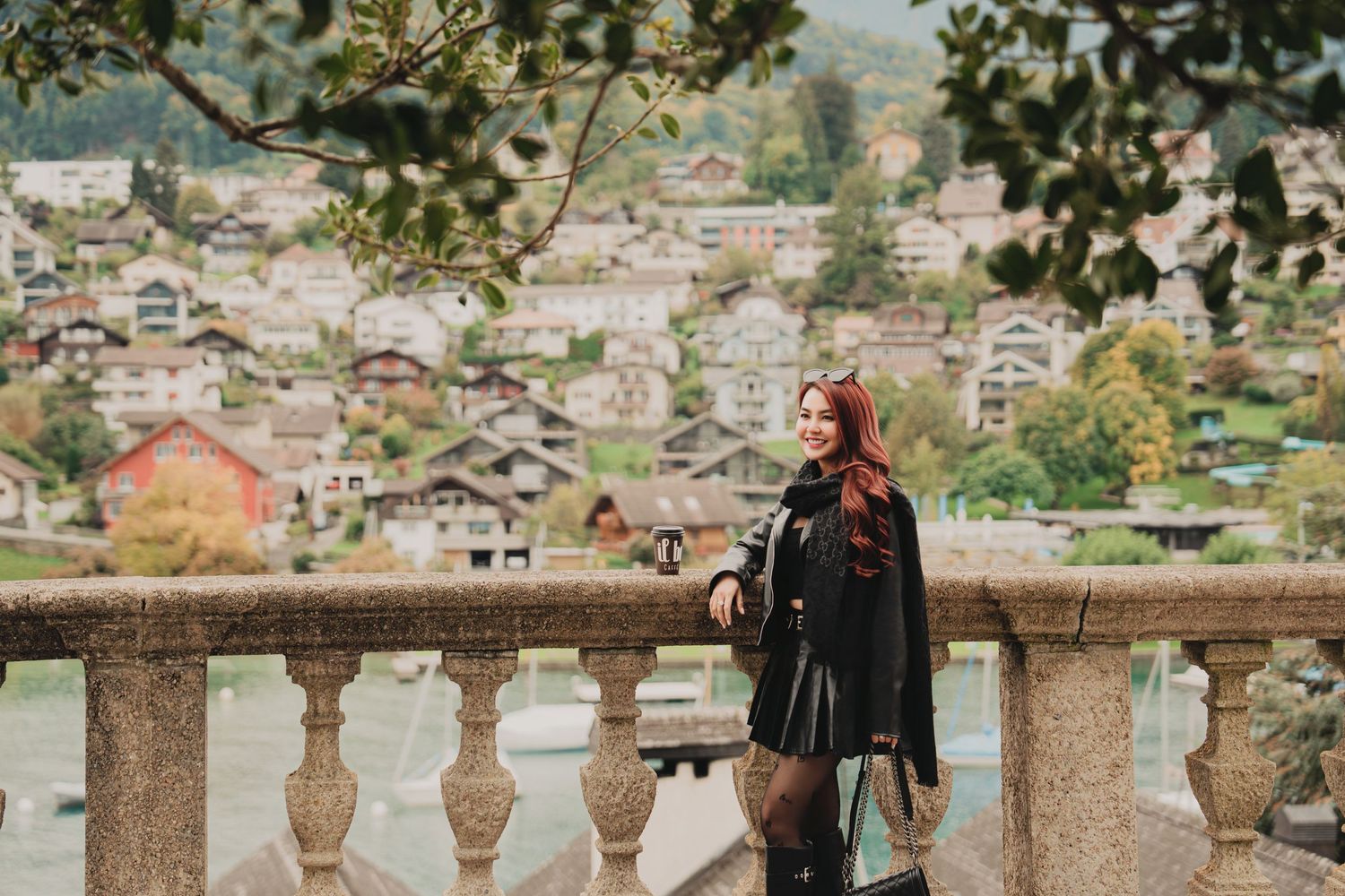 Elegant red-haired woman in black coat poses at stone balcony railing with scenic Swiss lakeside town in background.