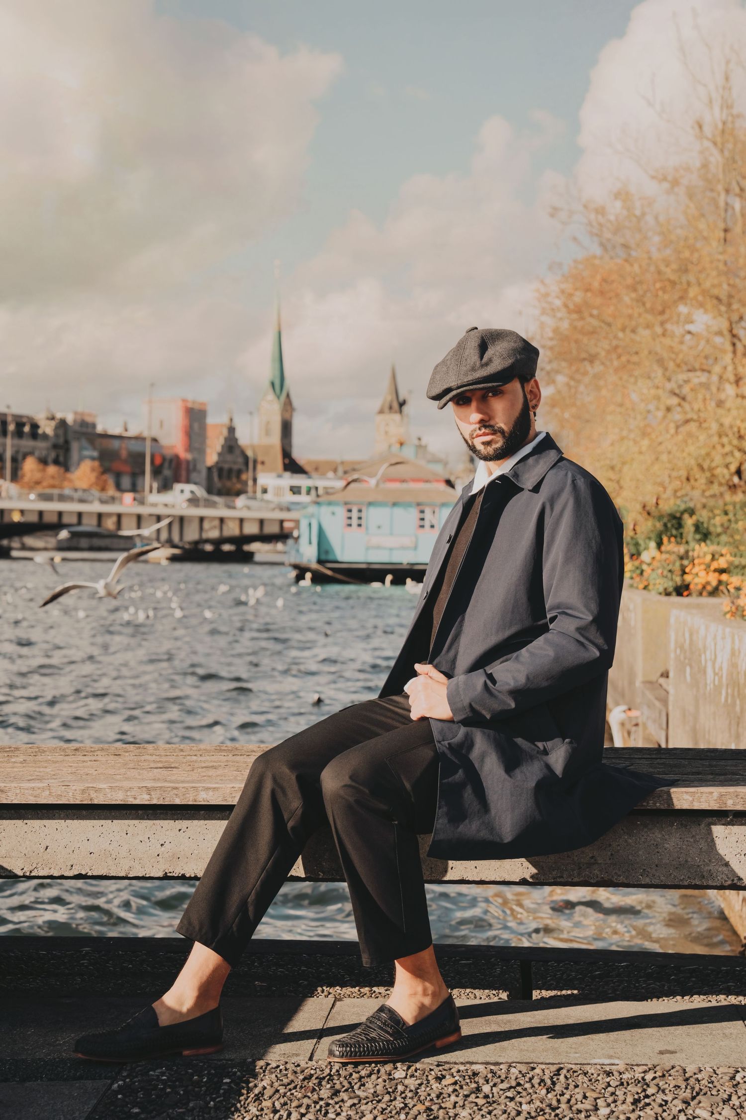 Bearded man in navy coat and flat cap sits on waterfront bench with church spires in background.