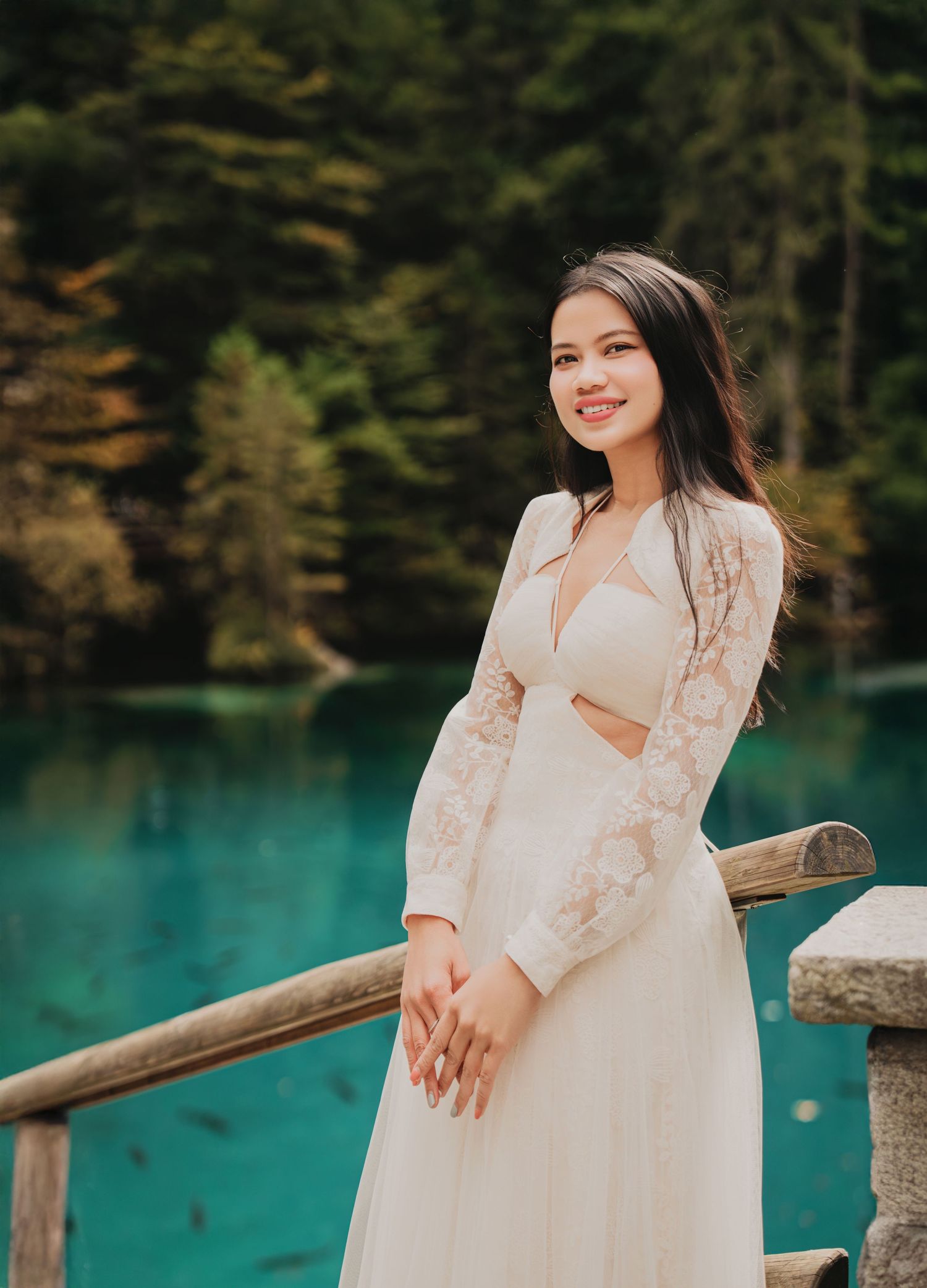 Smiling young woman in lace white dress poses by turquoise lake in forest setting.