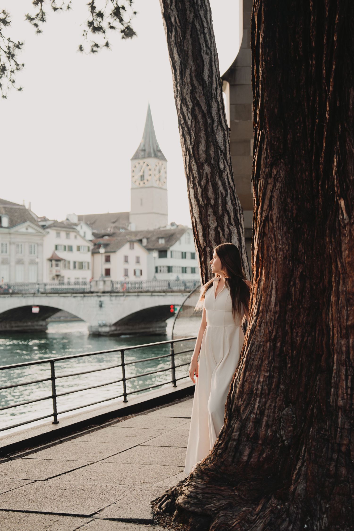 Woman in white dress standing beside large tree trunk along Zurich riverbank with church tower view.