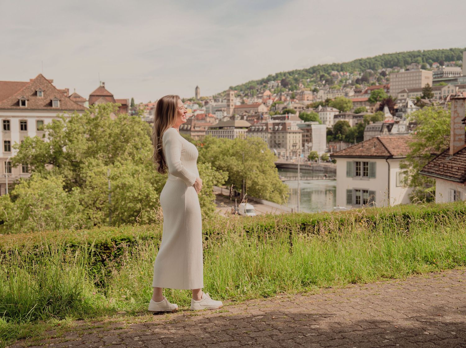 Pregnant woman in white dress gazing at European city skyline from elevated cobblestone path.
