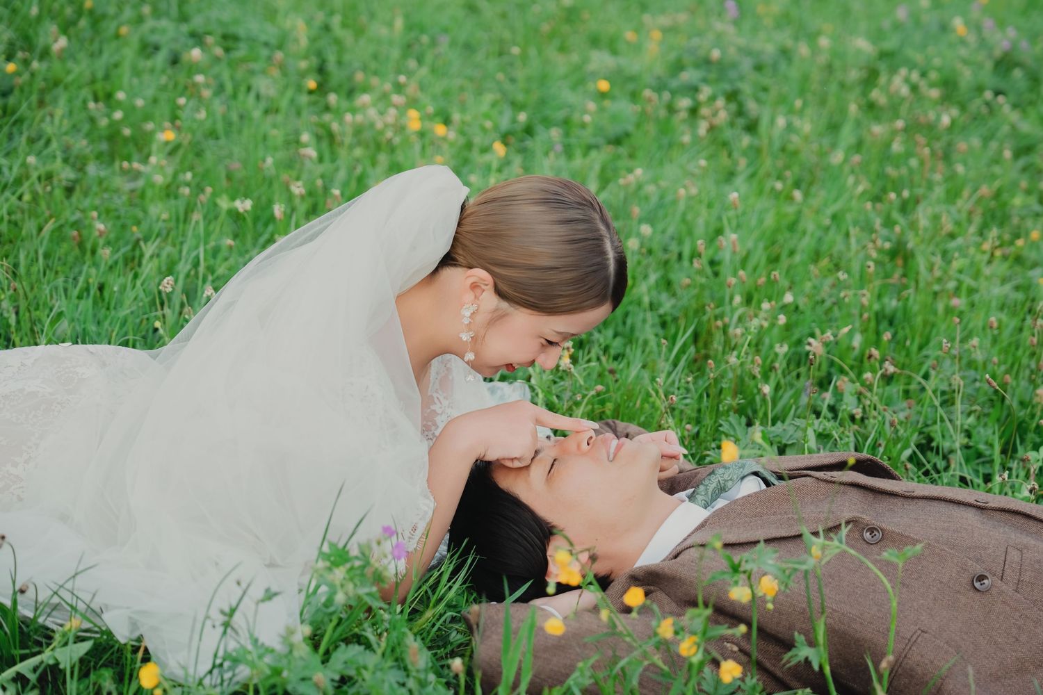 Bride leaning over groom lying in wildflower meadow during romantic alpine wedding shoot.