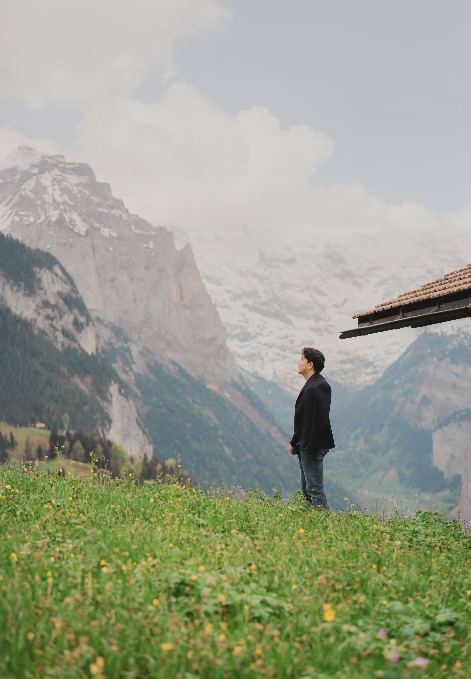 Man in black jacket standing on alpine meadow with snow-capped Swiss mountains and valley backdrop.