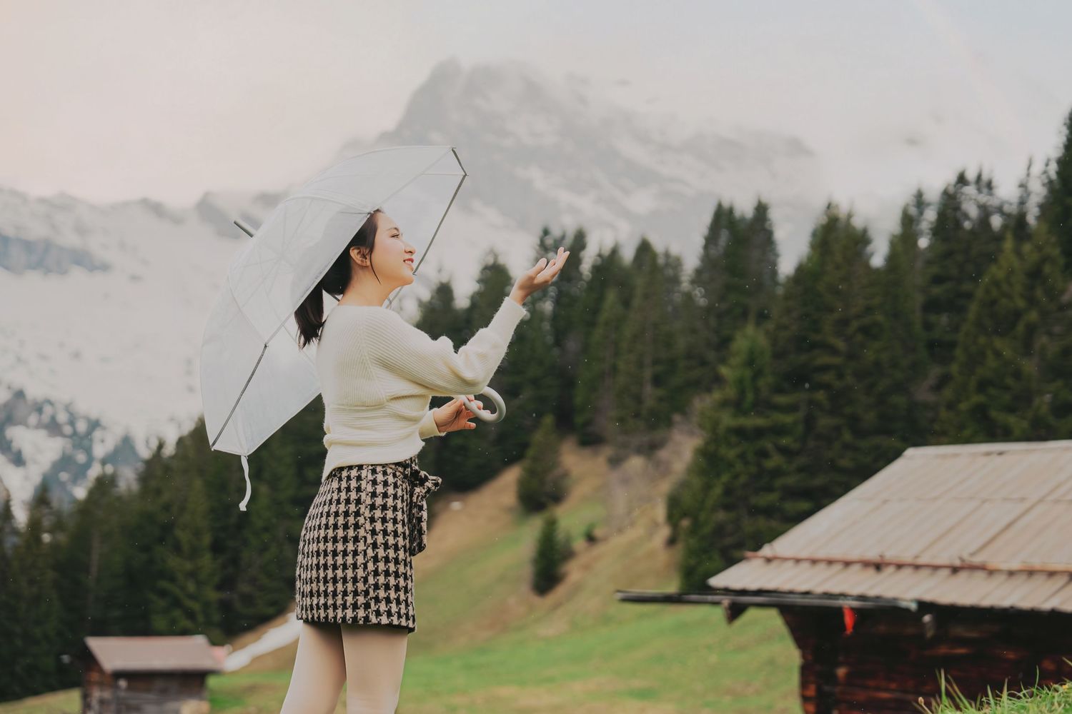Woman with umbrella checking for rain on alpine meadow with snow-capped mountains and pine trees in Switzerland.
