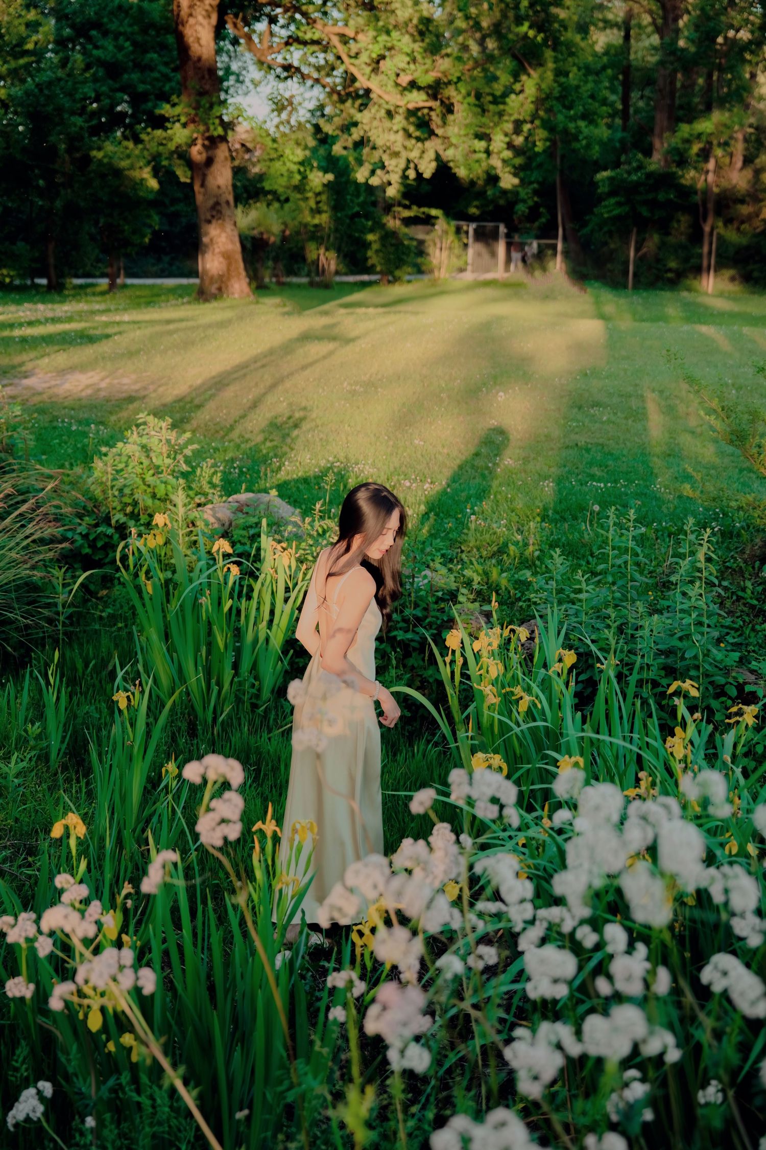 Woman in pink blouse and wide-leg pants standing among wildflowers in a lush green park at golden hour.
