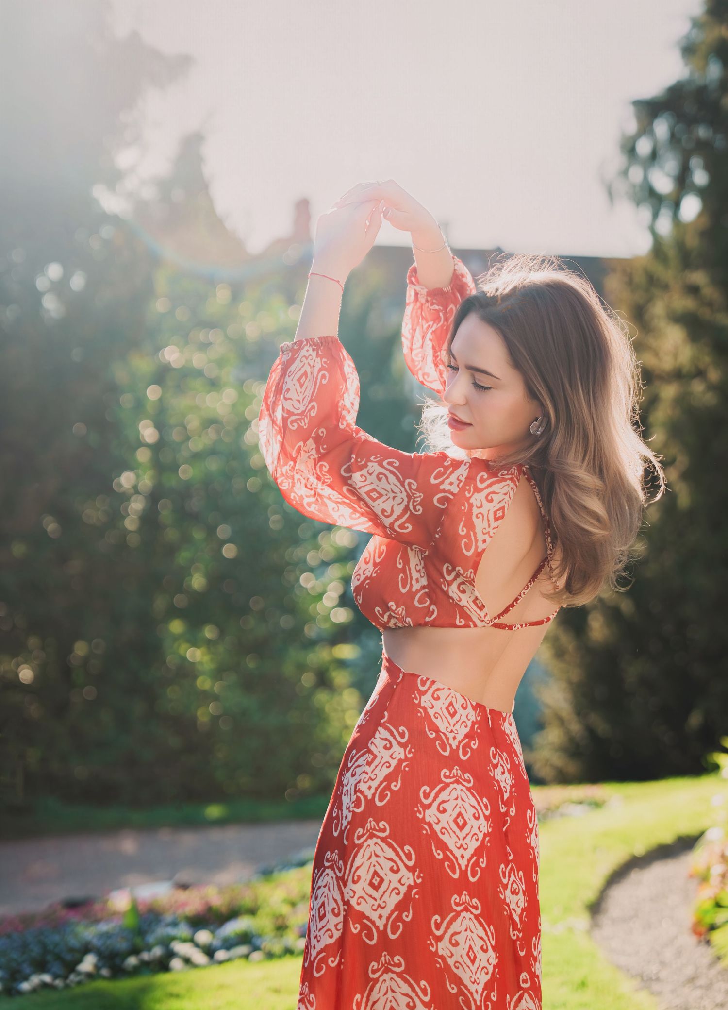 Woman in red boho print backless dress poses gracefully in a sunlit garden.