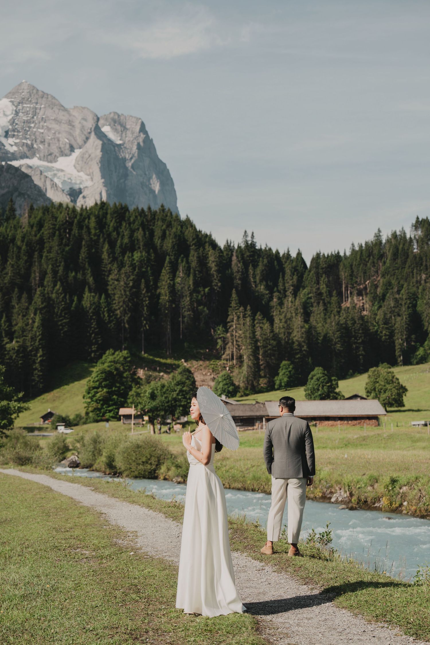 Bride with parasol and groom stroll beside glacial river in lush Swiss alpine valley setting.