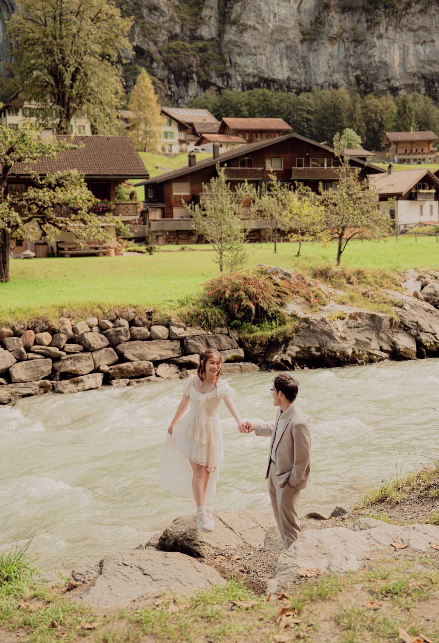 Couple holding hands on rocky riverbank with charming Swiss chalets and green meadows in Lauterbrunnen.