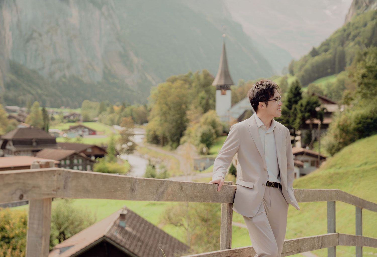 Stylish man in beige suit leaning on wooden fence with picturesque Swiss alpine village backdrop.