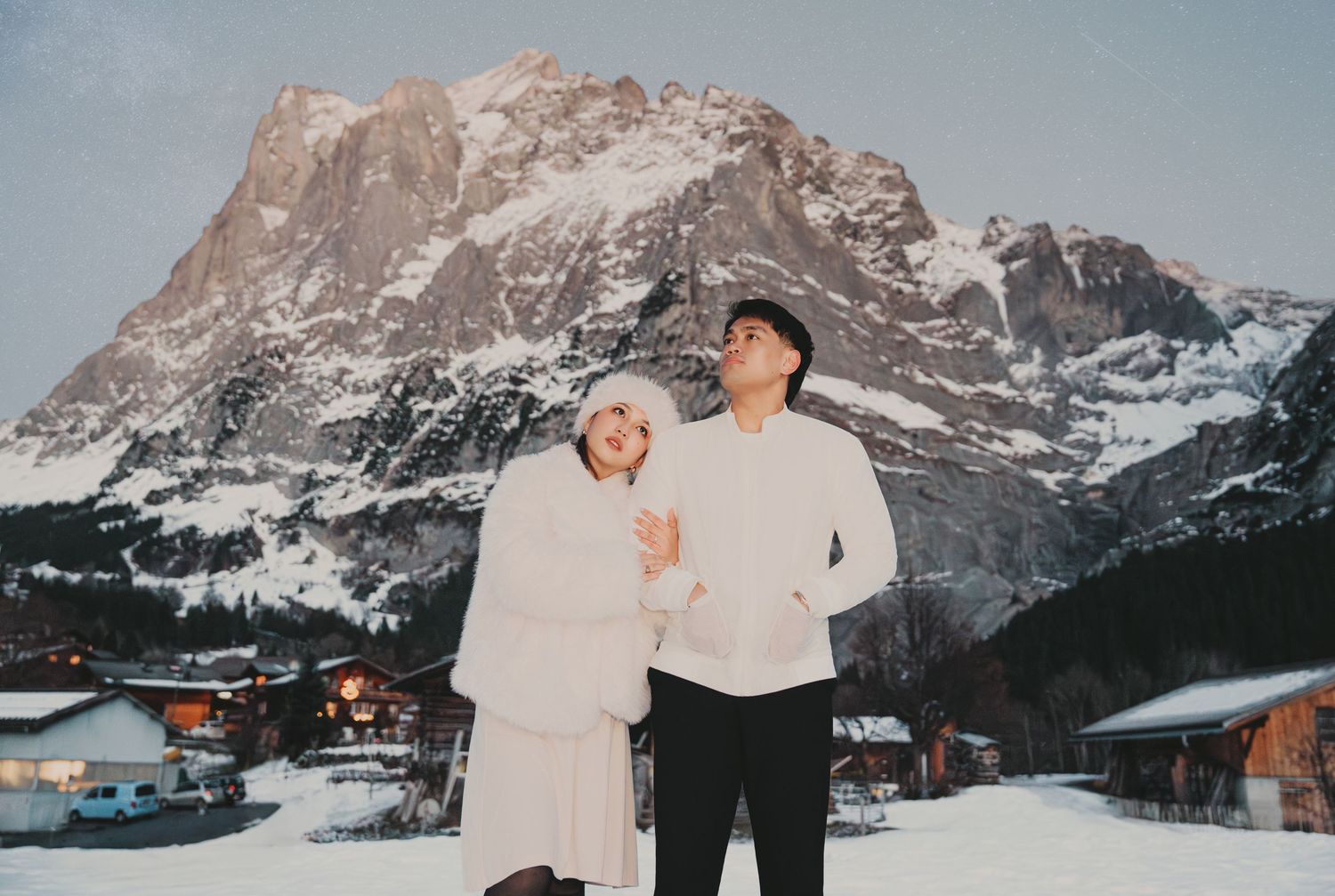 Couple in white outfits posing in snowy alpine village with dramatic mountain backdrop in winter.