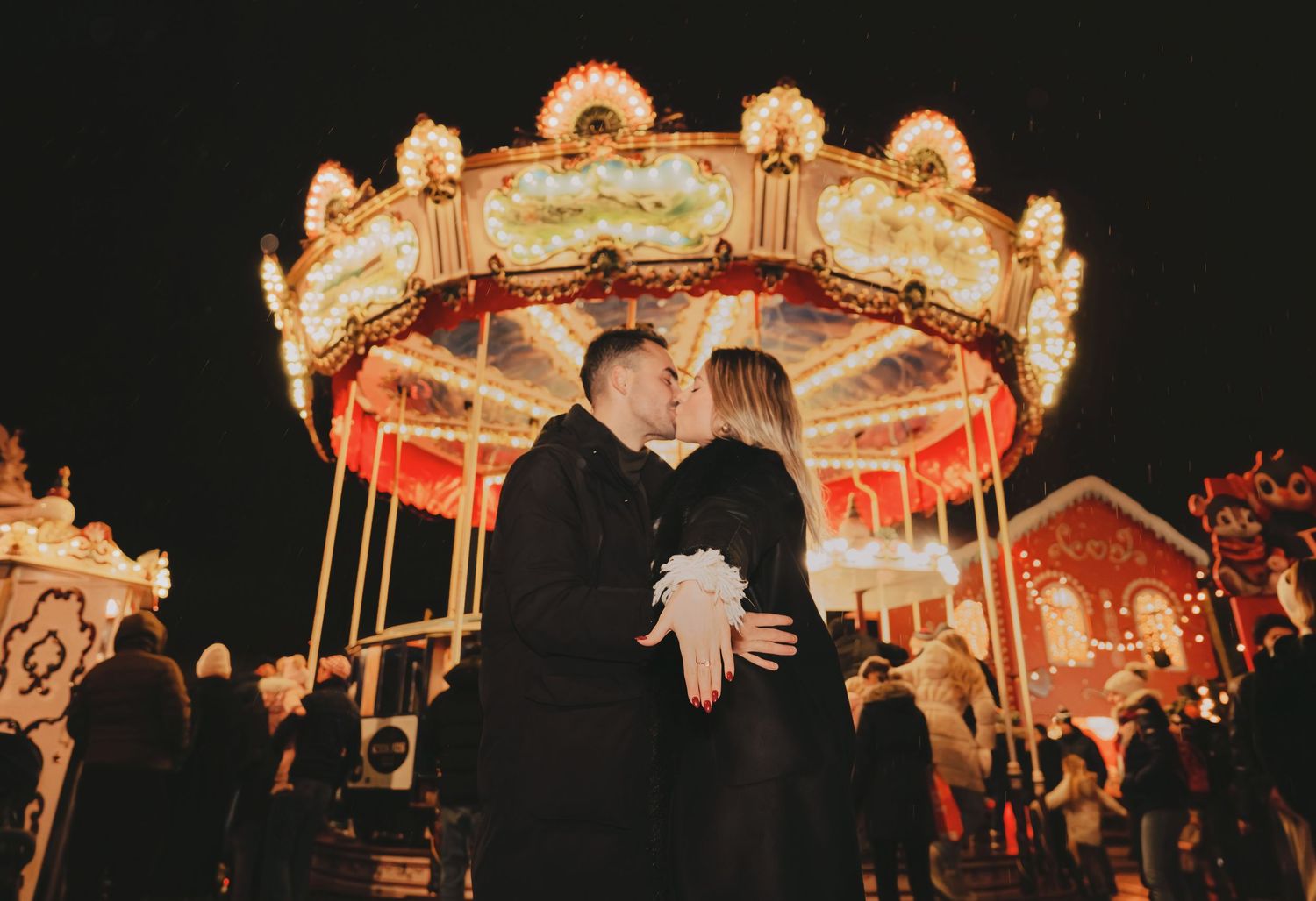 Couple sharing a romantic kiss in front of a glowing carousel at night.