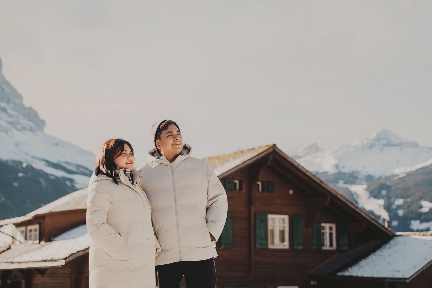 Couple in matching white winter coats standing together in snowy alpine village with mountain backdrop.