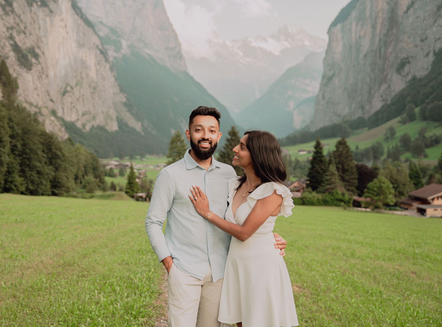Affectionate couple embracing on a green meadow with a dramatic Swiss mountain valley in the background.