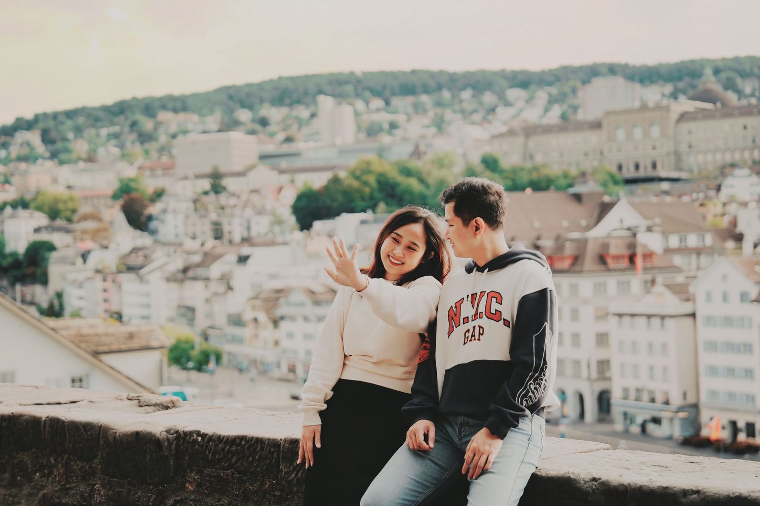 Happy couple sitting on stone wall admiring panoramic view of Zurich city skyline together.