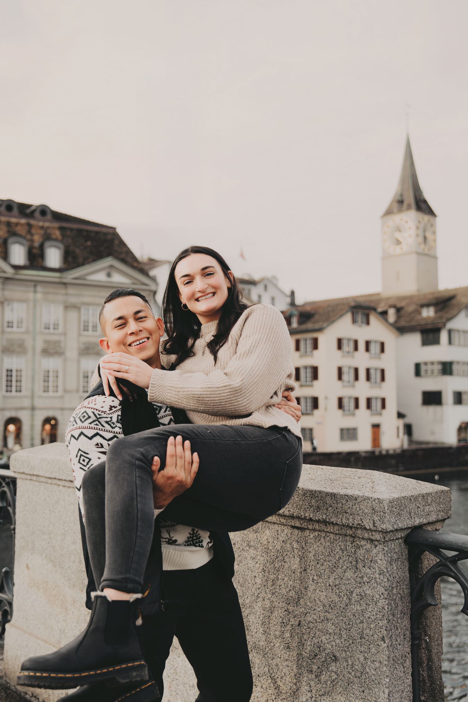 Happy couple posing on a stone bridge railing with Zurich's iconic St. Peter's Church tower in the background.