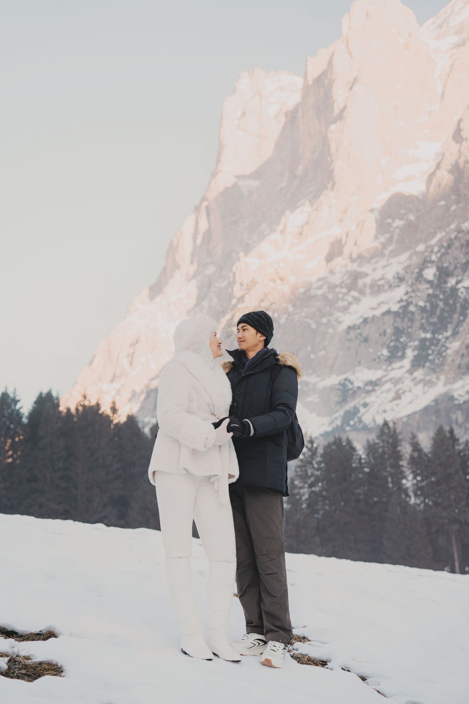 Couple standing together in snowy mountain valley with pine trees and rocky peaks at sunset.