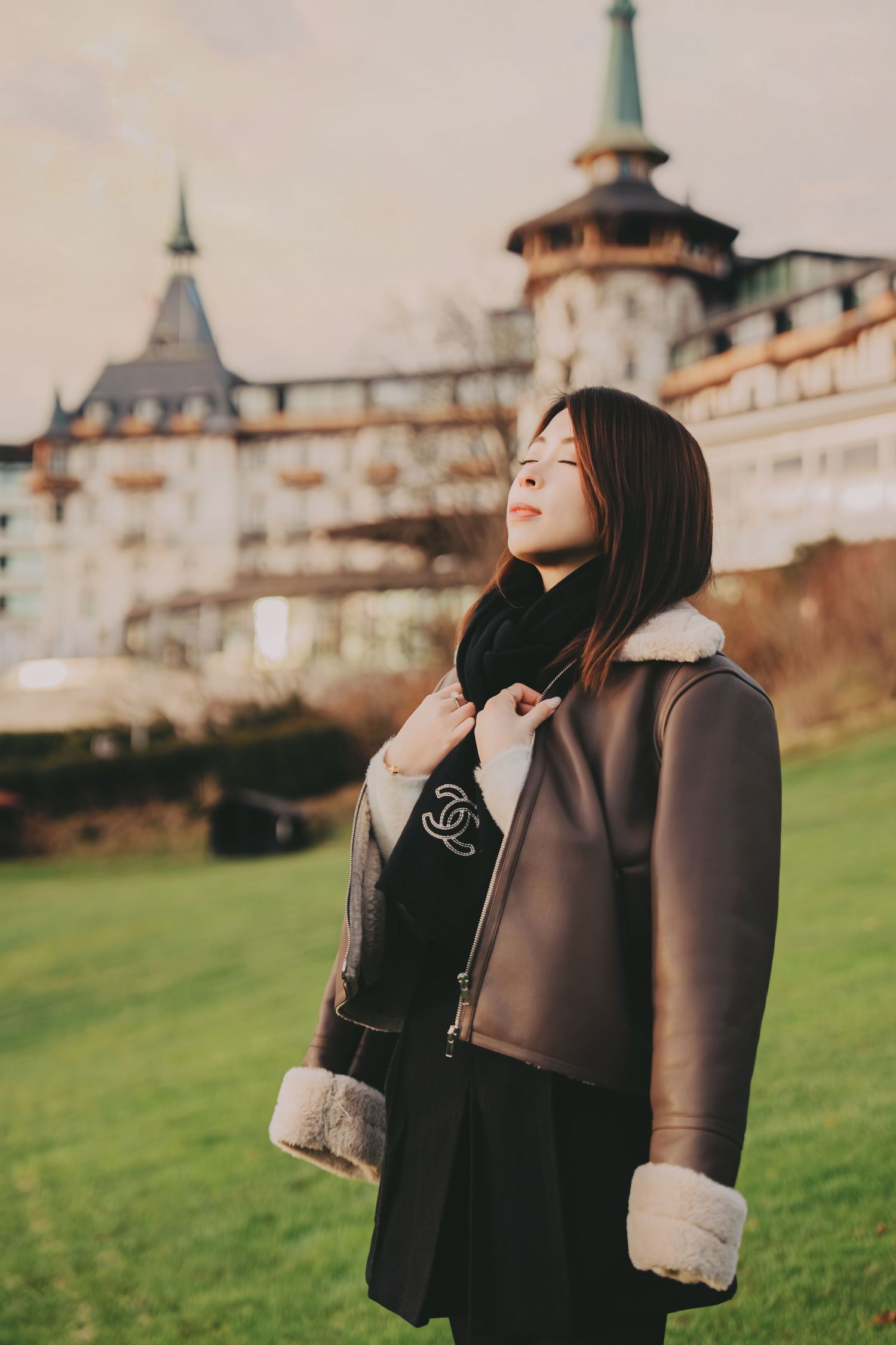 Young Asian woman in brown shearling coat smiling with eyes closed before European château.