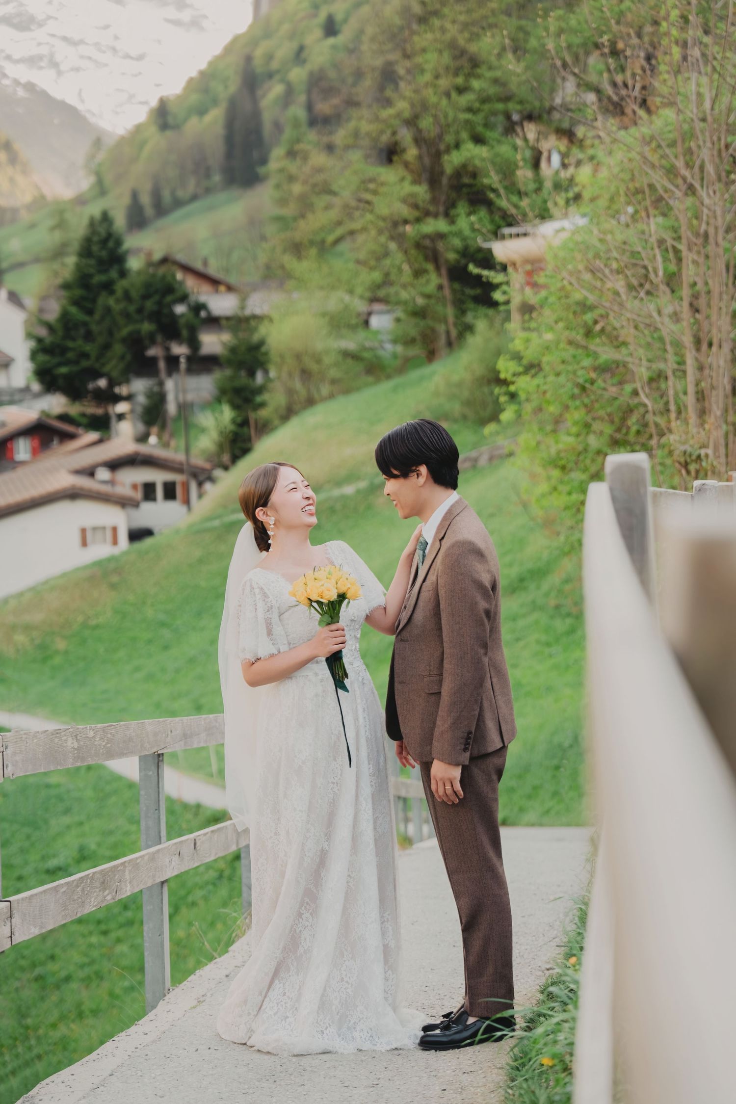 Smiling bride holding yellow flowers as groom in brown suit gazes at her on a bridge in Switzerland.