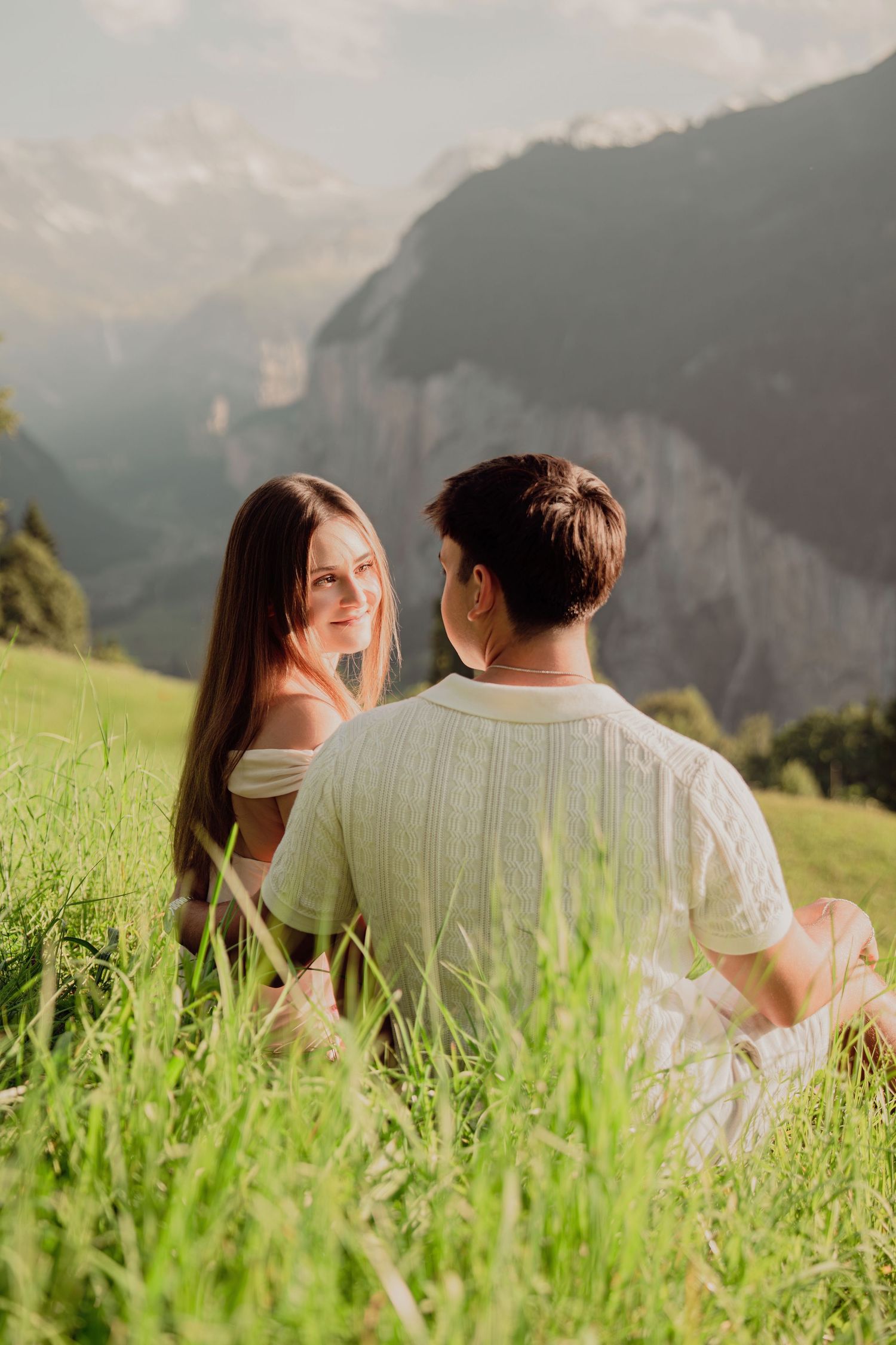 Smiling woman sitting with partner in green alpine meadow with dramatic Swiss mountain scenery.