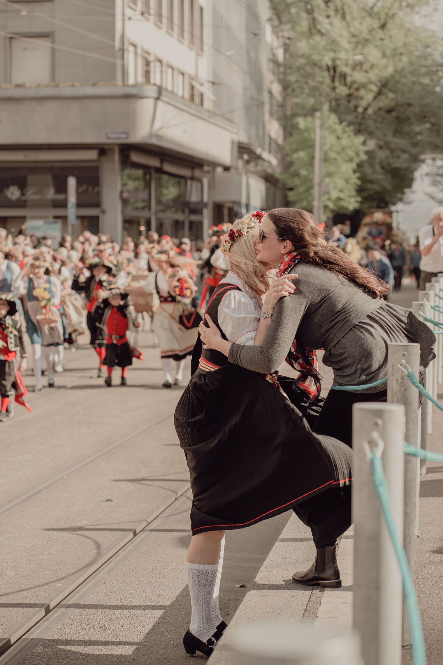 Two women in traditional Swiss dirndl costumes embracing warmly while watching a colorful folk parade on a city street.