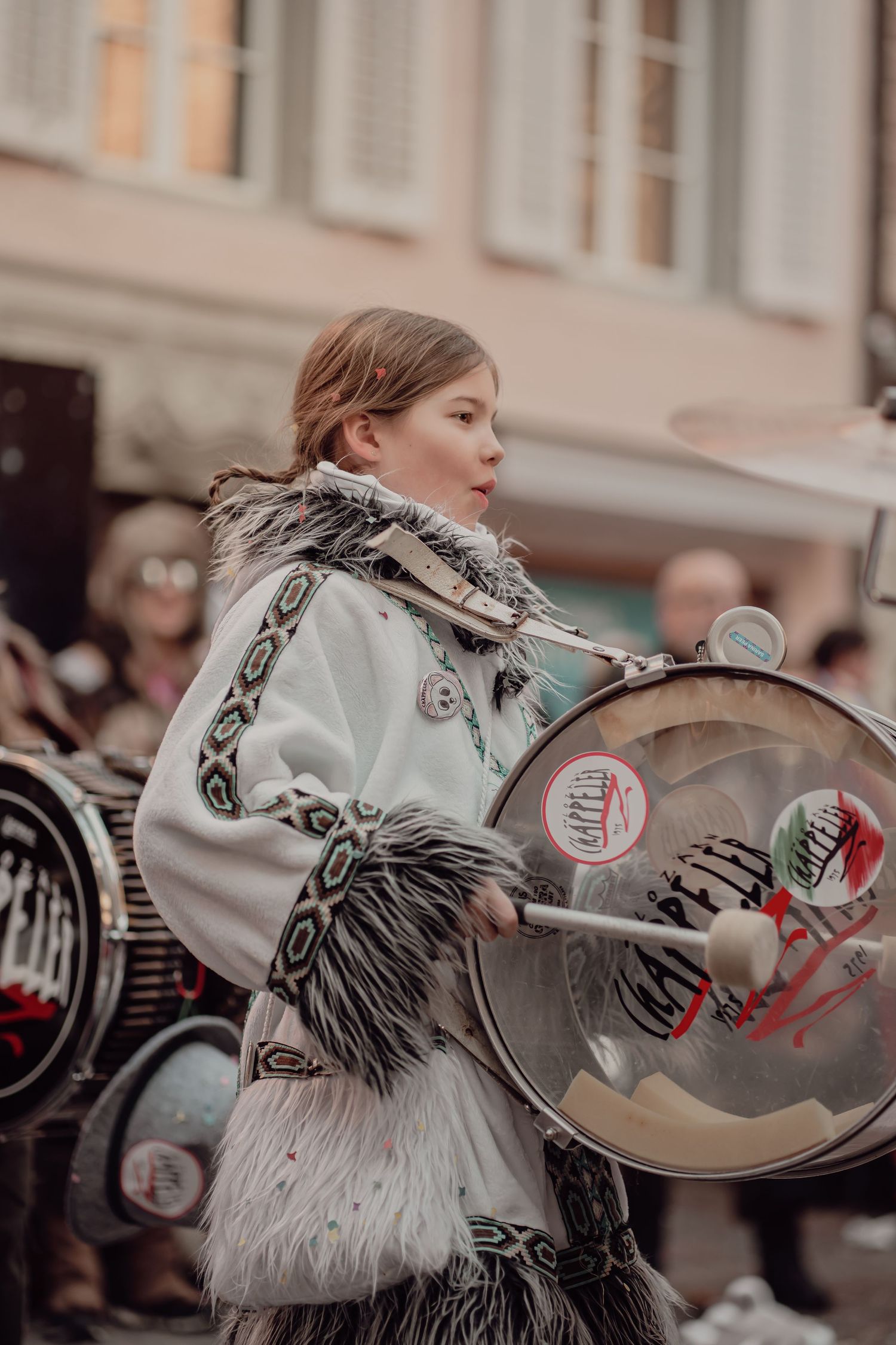 Young girl in ornate white fringed costume playing a large bass drum in a street parade.