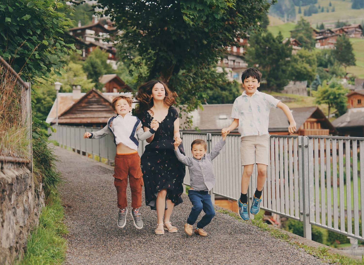A woman and three children joyfully run along a scenic Swiss village path surrounded by lush greenery.