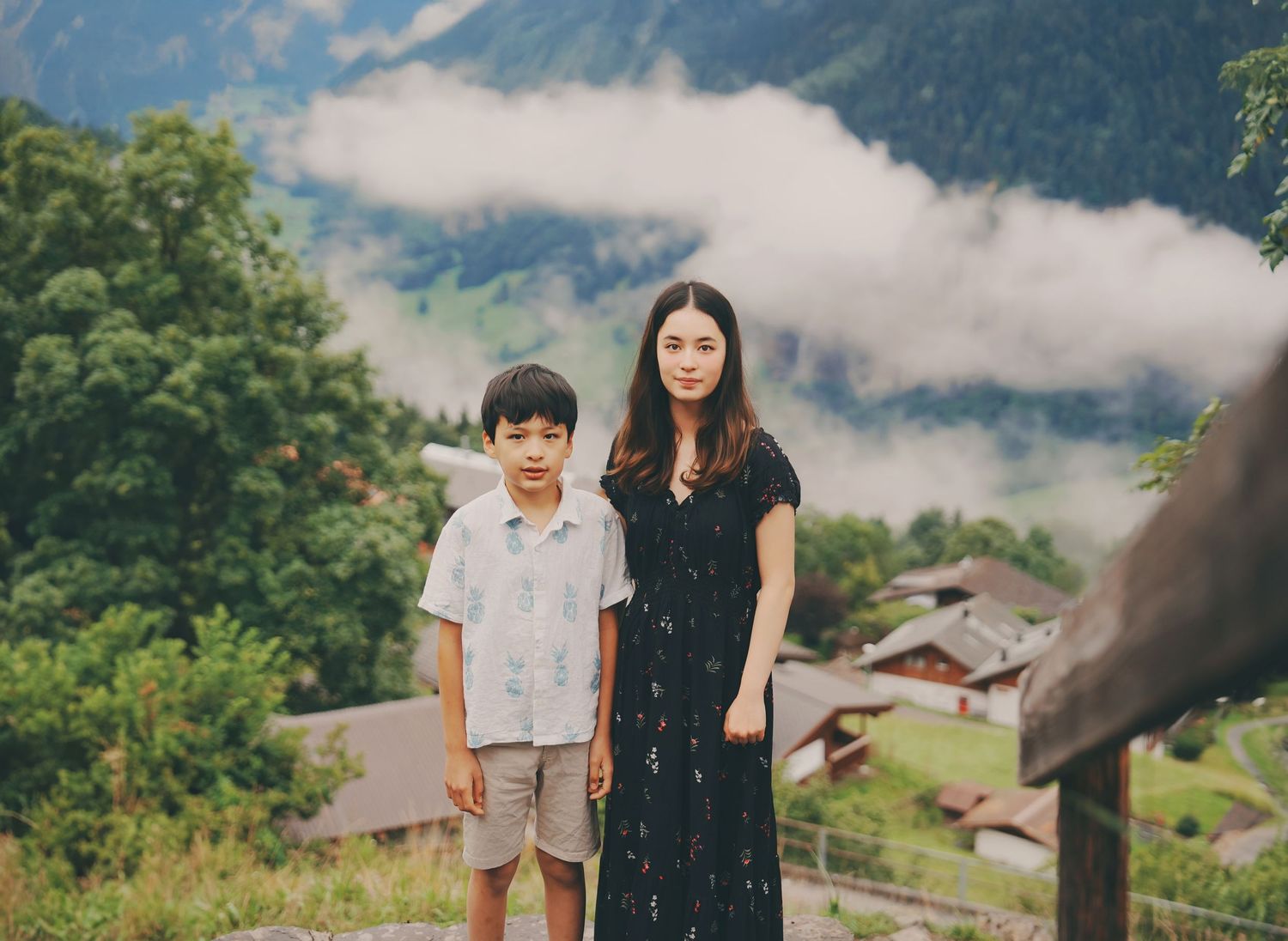 Mother in black dress and young son standing together with lush green Swiss mountain village in the background.