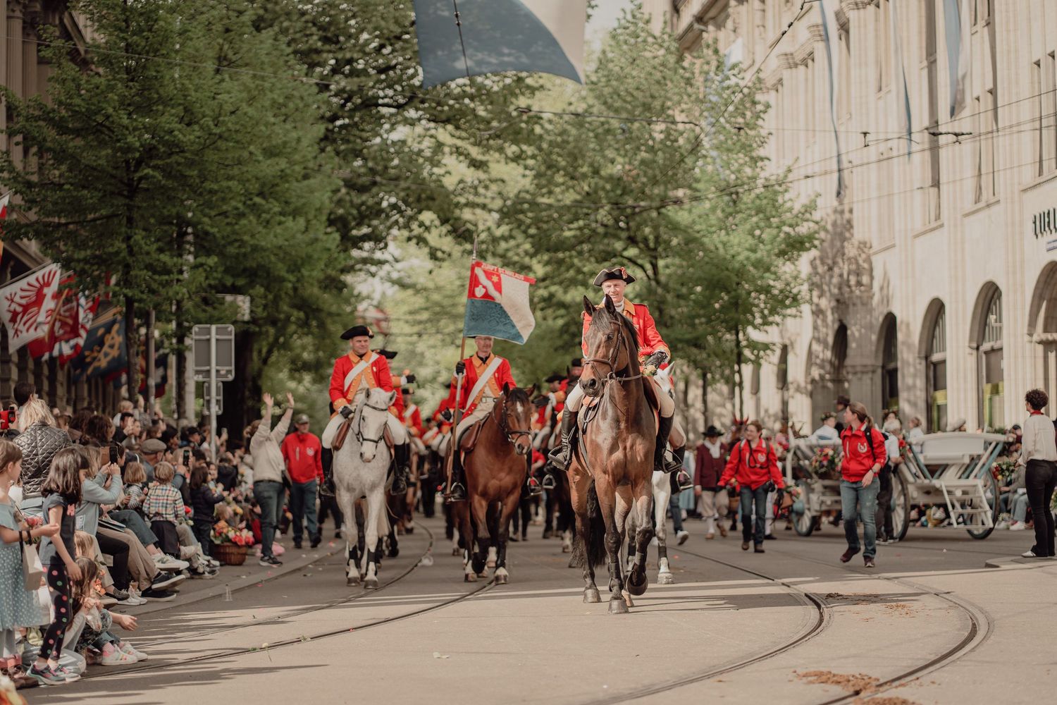 Riders in red historical uniforms on horseback lead a colorful street parade through a European city.