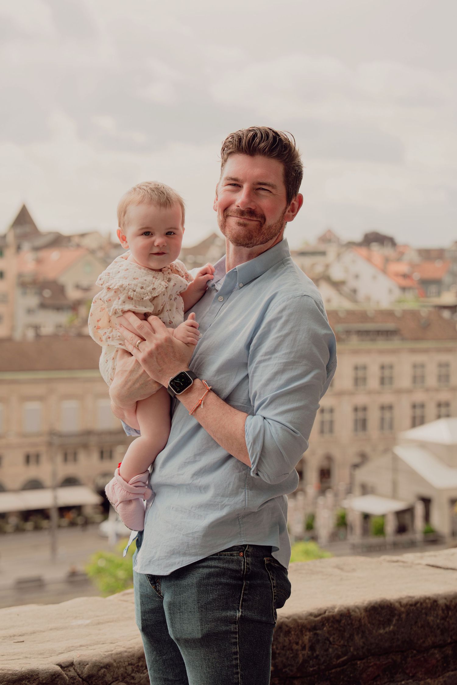 Smiling father holding baby girl with historic European city in background.