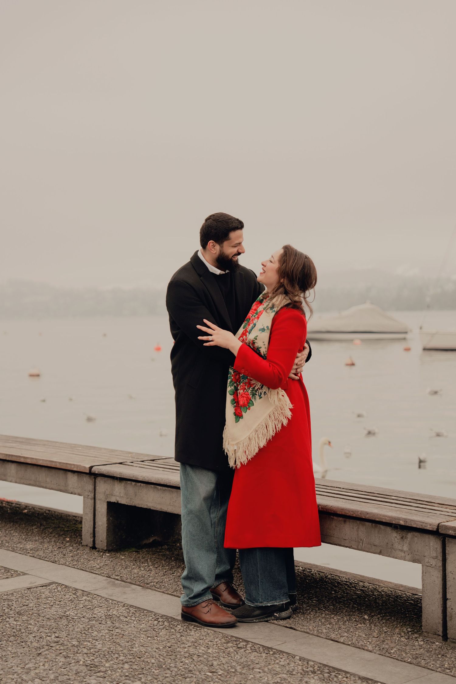 Couple embracing and gazing at each other by a misty lakeside, woman in red coat with floral scarf.