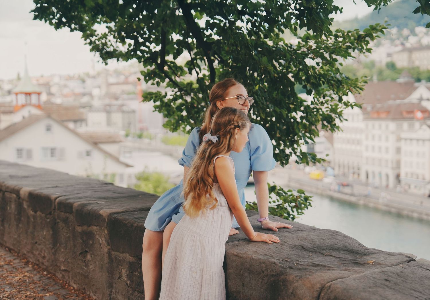Two girls leaning over a stone wall overlooking a scenic European cityscape.