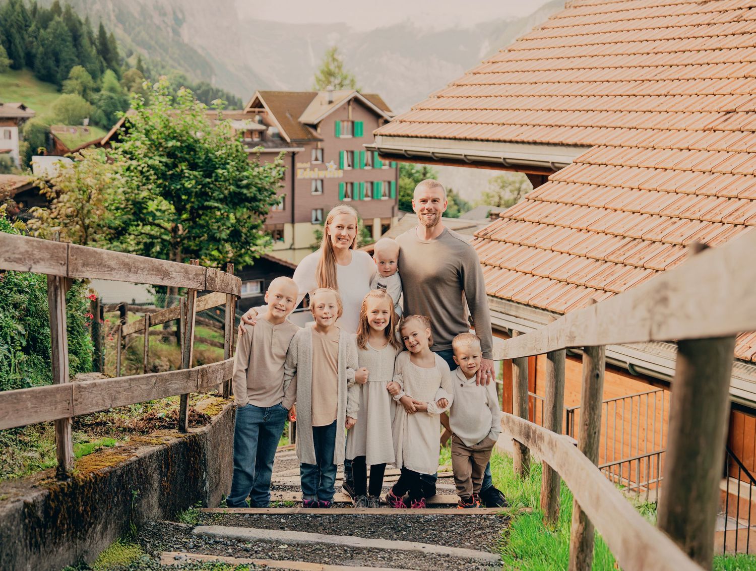 Family of seven with parents and five children smiling on outdoor steps in a scenic Swiss mountain village.