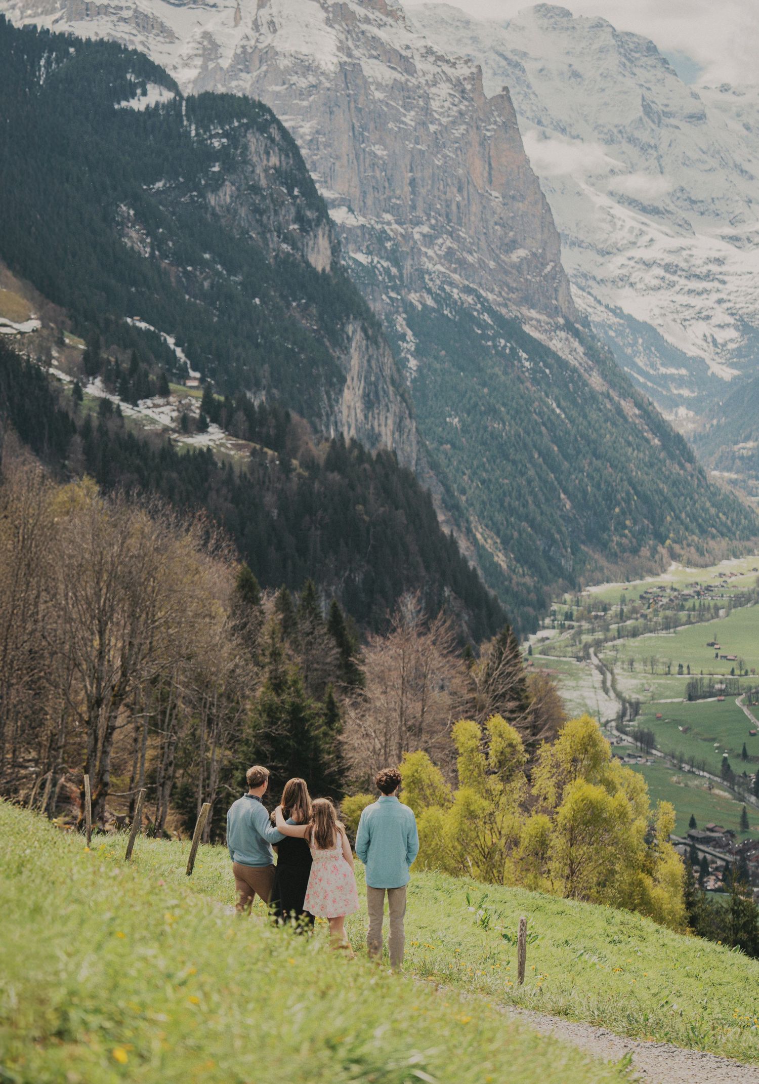 A family of four walks along a grassy hillside trail overlooking a stunning Swiss alpine valley below.