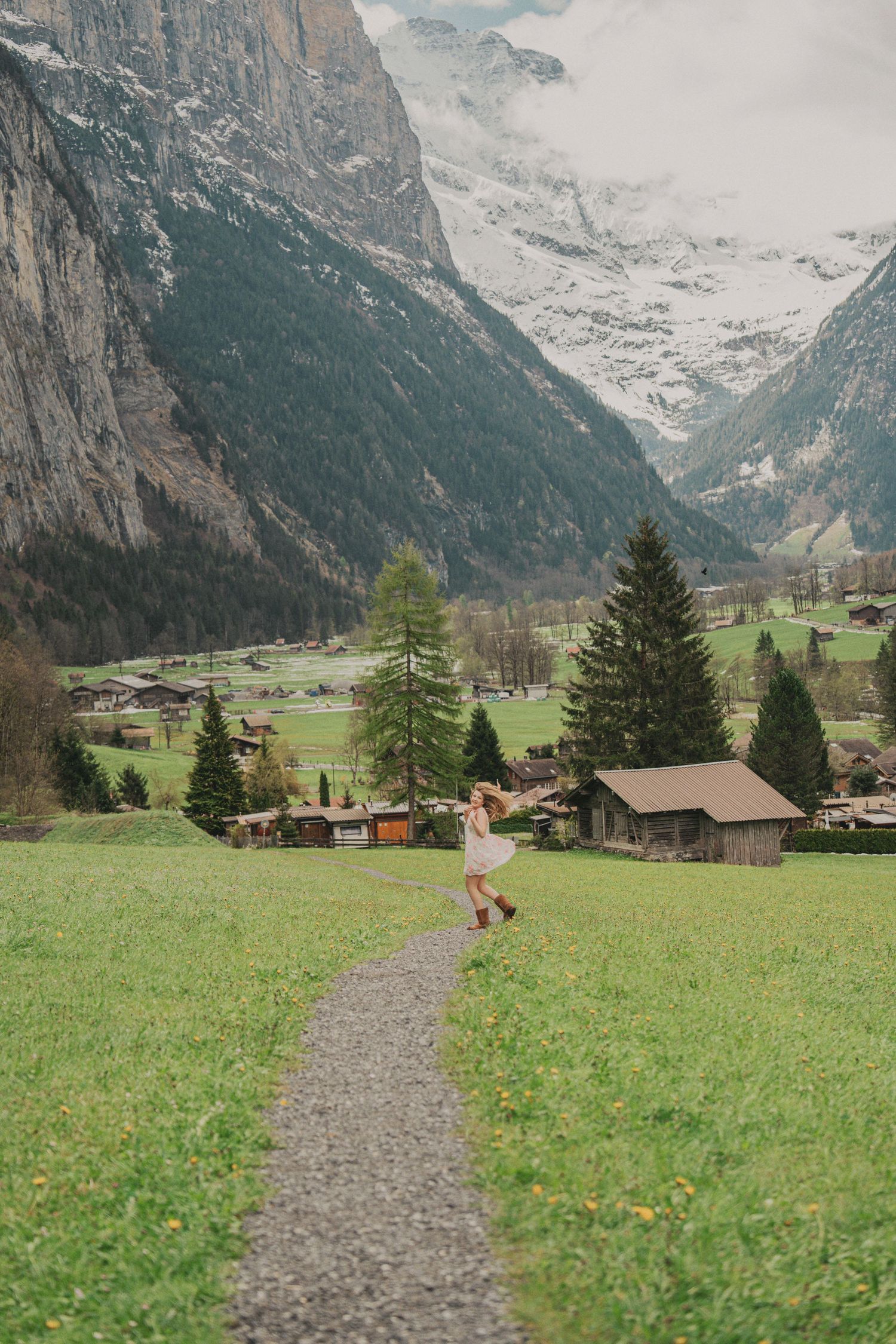Woman in floral dress walking on gravel path through lush green Swiss mountain valley village.