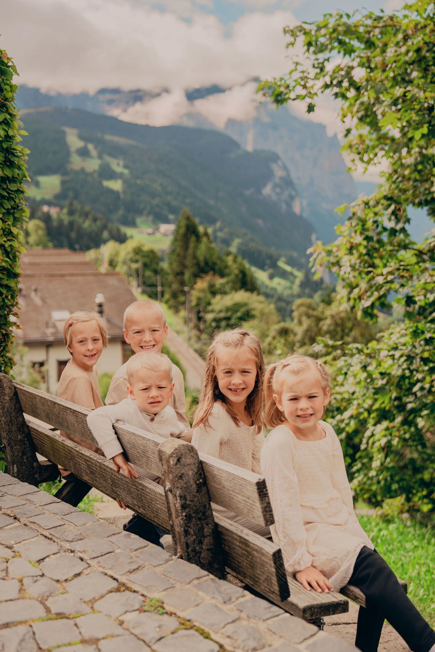 Five young children sitting and posing on a rustic wooden bench with scenic Swiss Alpine mountains in the background.