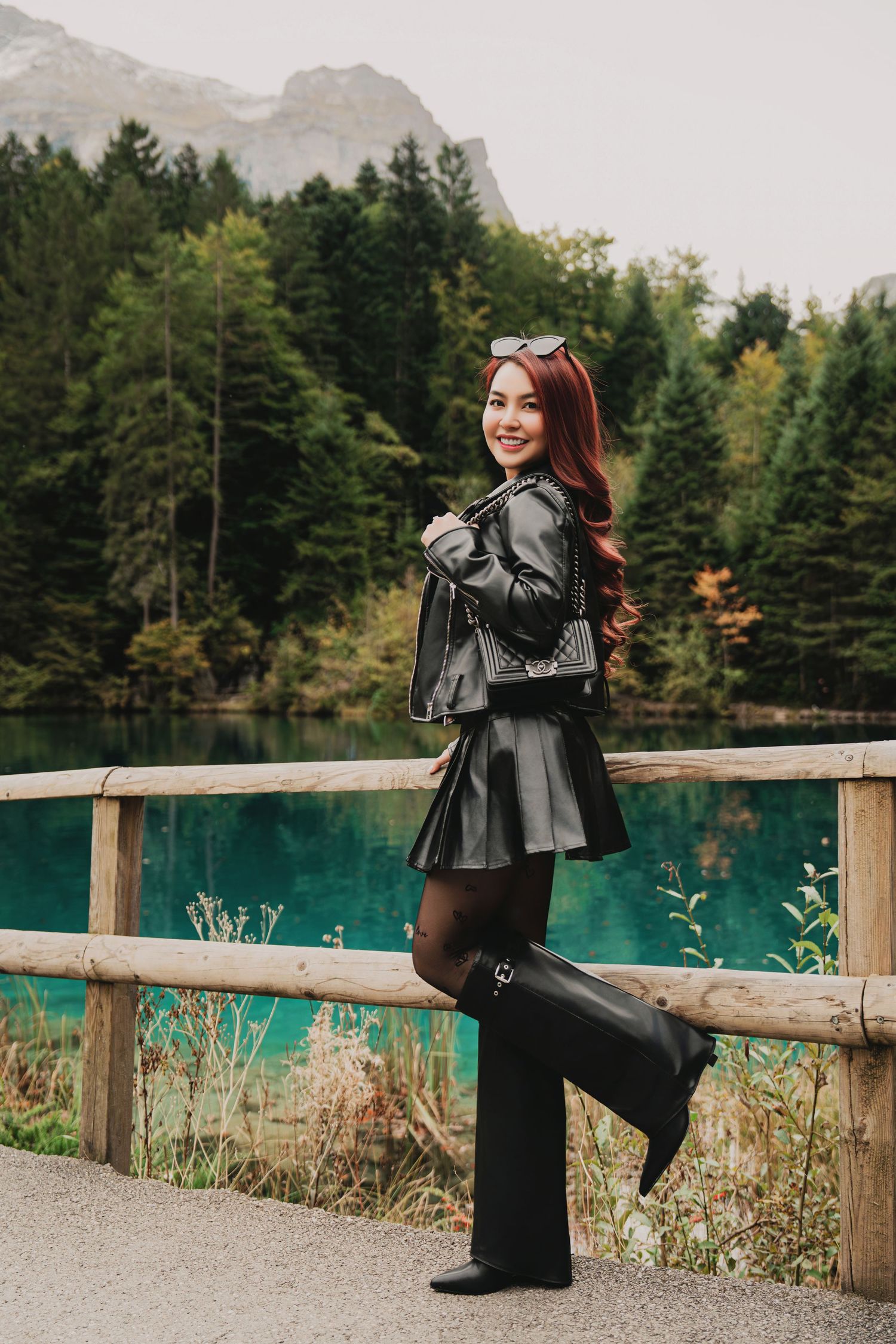 Smiling woman in black leather jacket holding bag beside vivid turquoise alpine lake and mountains.