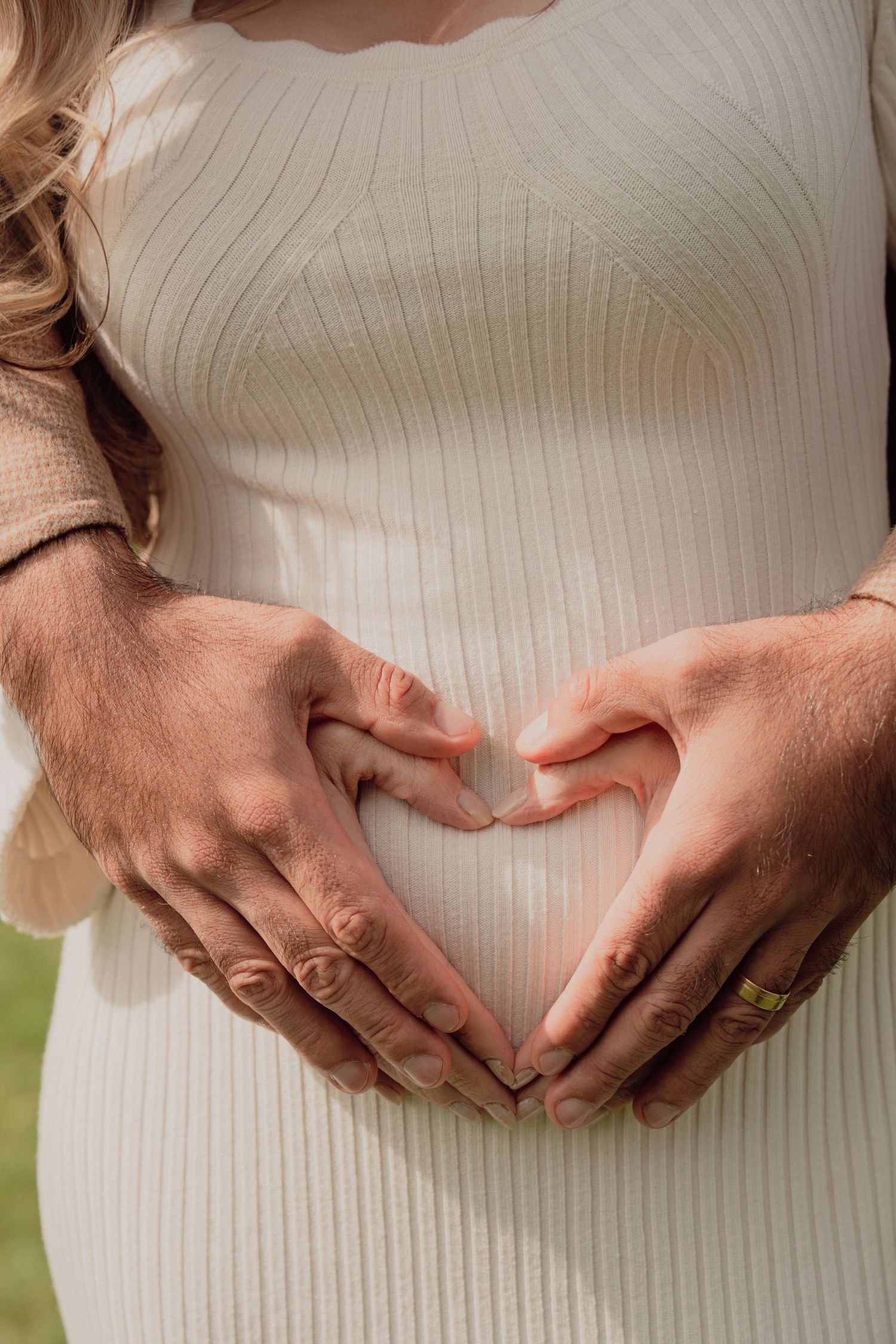 Close-up of couple forming a heart shape with their hands over a pregnant belly in white dress.